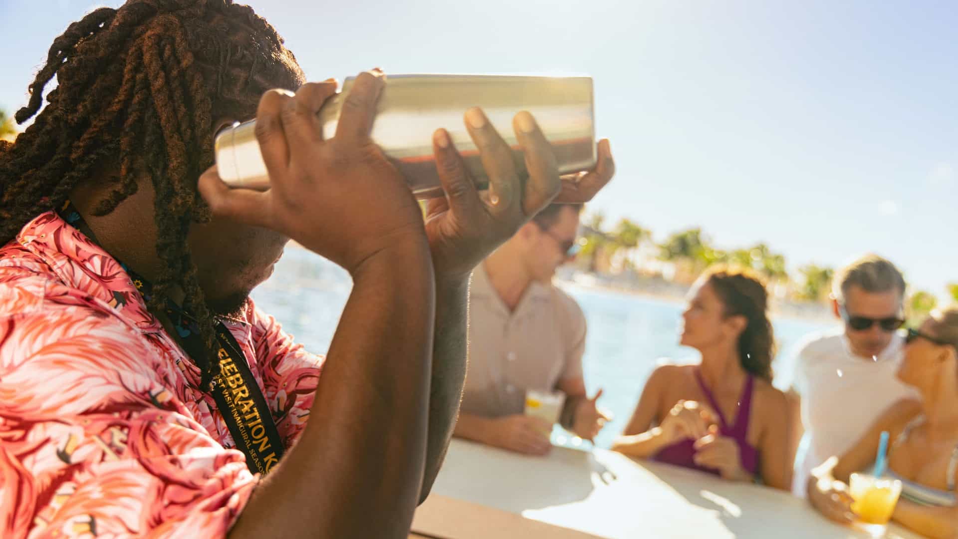 A bartender in a pink patterned shirt shakes a cocktail mixer at a sunny outdoor bar while guests relax and enjoy drinks in the background.