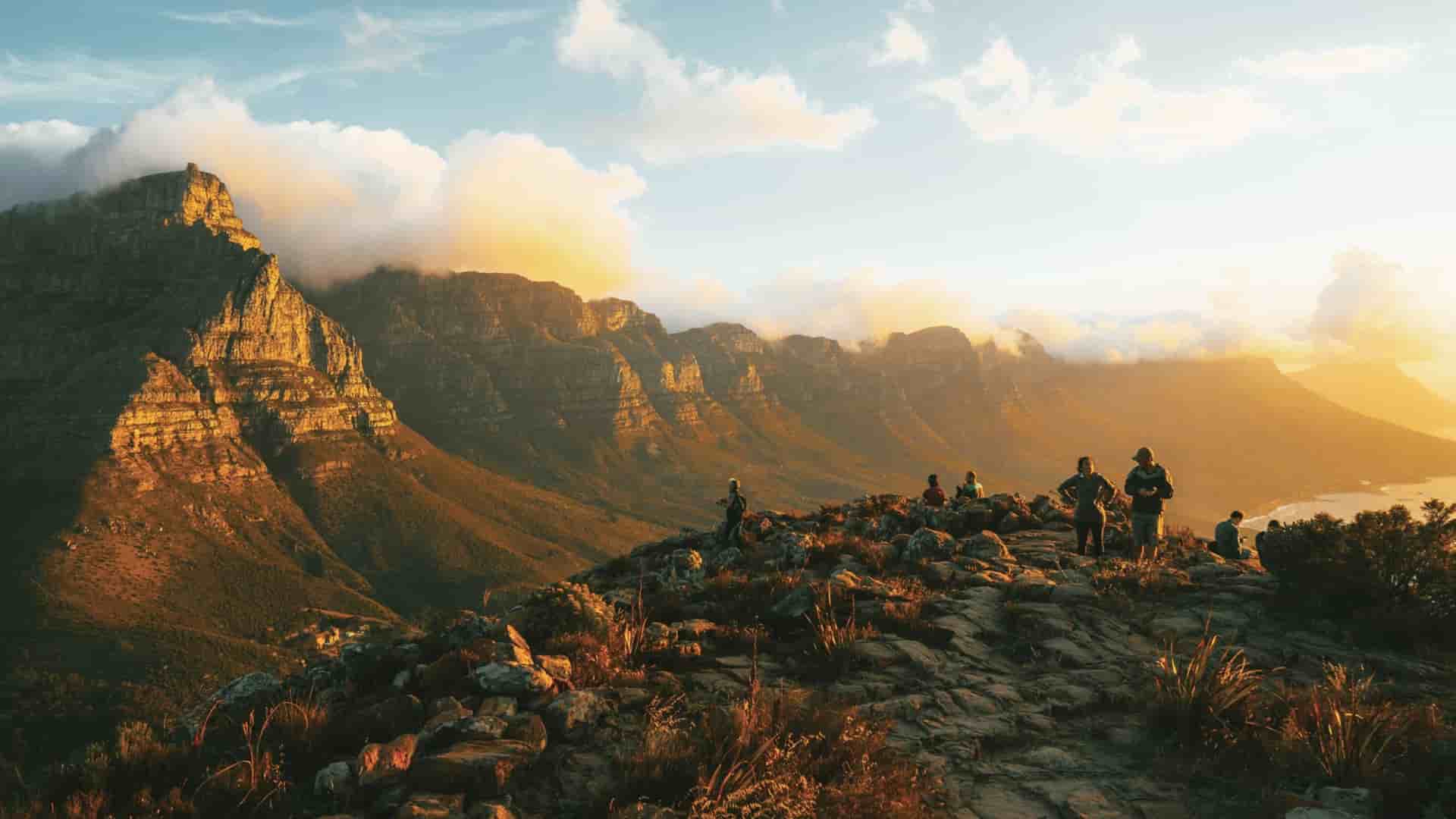 Hikers on the rocky trail at Lion's Head, Cape Town, with the Twelve Apostles mountain range glowing in golden sunset light.