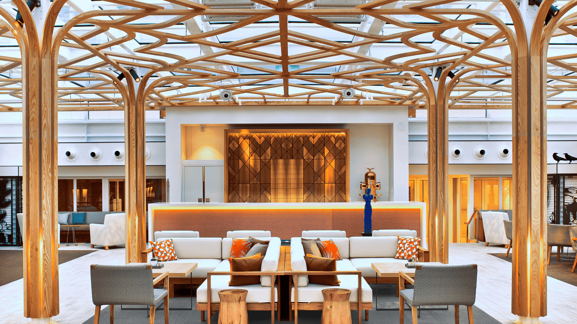 An elegant indoor lounge called the Wintergarden, featuring light wood columns that branch out into an intricate geometric lattice across a glass ceiling. The space is filled with modern white sofas, orange accent pillows, and a sleek bar in the background under bright, natural light.