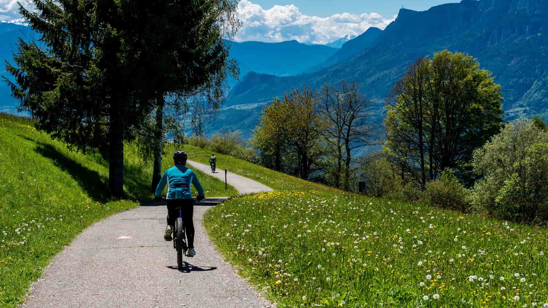 A wide shot image of a bicyclist in the foreground on tarmac along a range of mountains and greenery withe blue skies and clouds. In the background is another bicyclist further away into the distance.