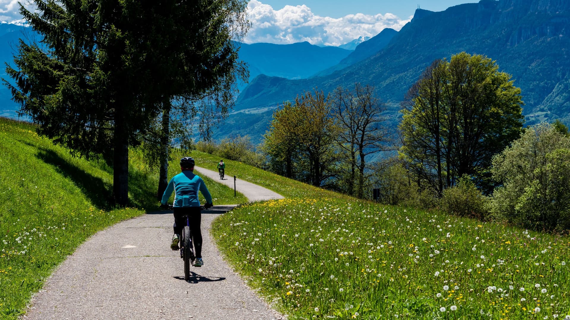 A wide shot image of a bicyclist in the foreground on tarmac along a range of mountains and greenery withe blue skies and clouds. In the background is another bicyclist further away into the distance.