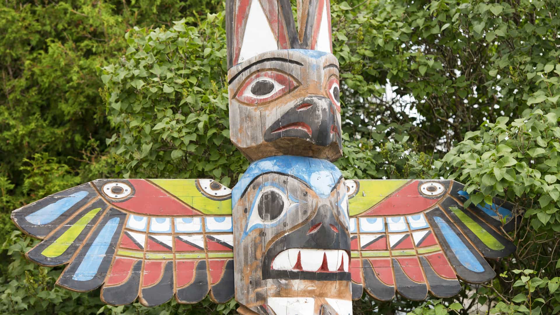 Detailed close-up of a traditional hand-carved and painted Native Alaskan totem pole, featuring animal and human figures, set against a backdrop of green trees.