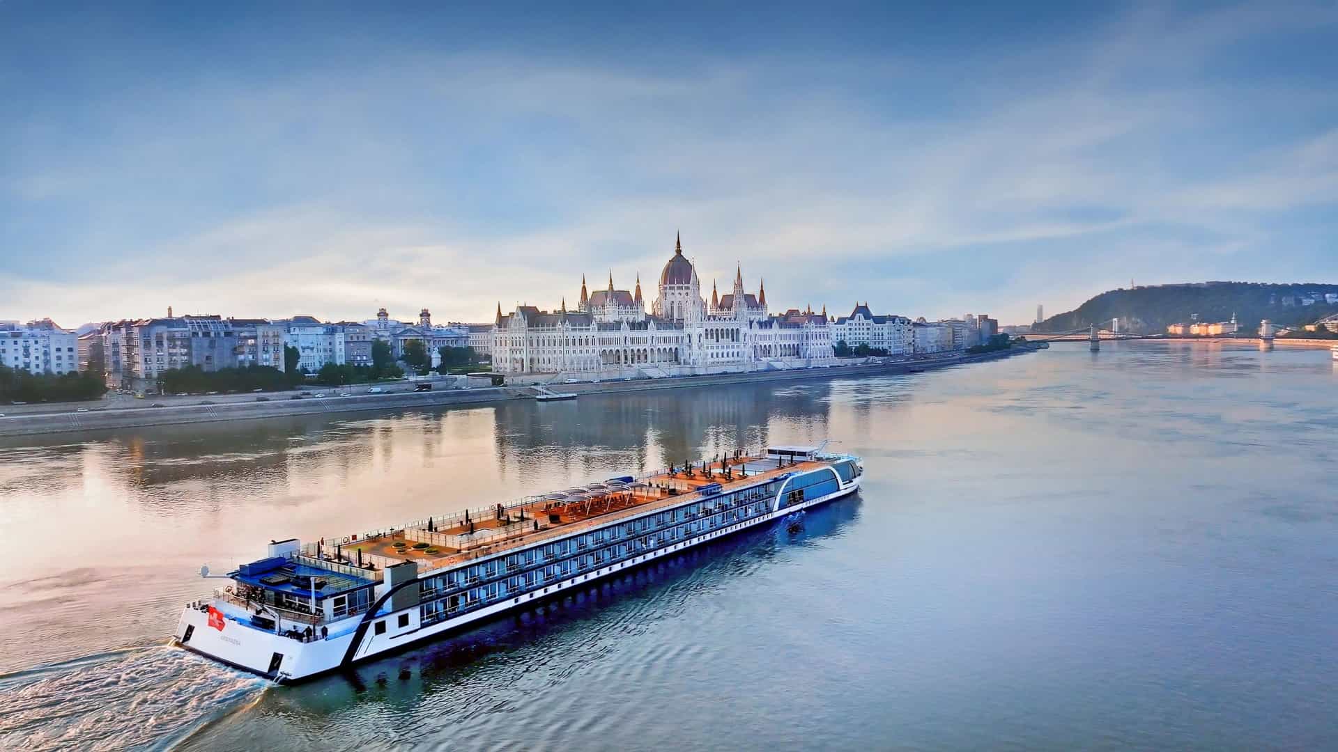 The AmaWaterways AmaMagna luxury river cruise ship sailing the Danube River with the iconic Hungarian Parliament Building in Budapest, Hungary, in the background.