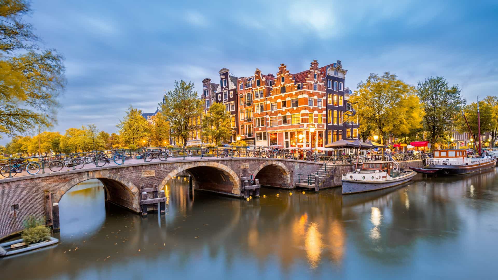 AmaWaterways Northern Europe river cruise destination in Amsterdam, Netherlands, showcasing a picturesque canal with historic gabled houses, an arched bridge adorned with bicycles, and glowing streetlights at dusk.