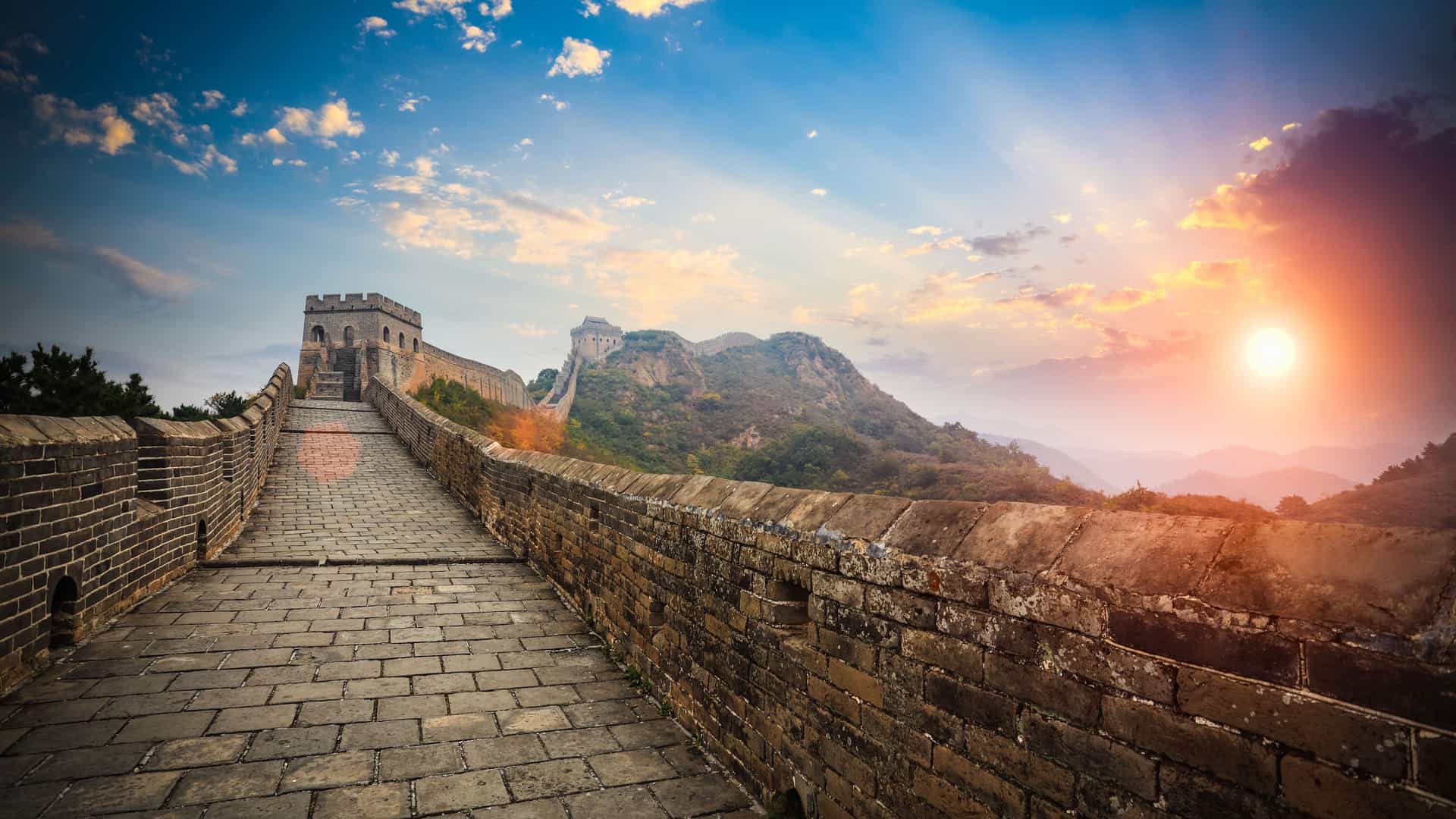 An iconic view of the Great Wall of China at sunset, specifically the Jinshanling section in China. The stone path of the wall, with its protective brick barrier, recedes into the distance over rolling hills and mountains. A traditional watchtower stands tall in the middle ground, and another is visible on a distant peak. The sky is a dramatic mix of brilliant blue and fiery orange and yellow from the setting sun, with rays of light illuminating the scene, highlighting this famous Asian landmark.