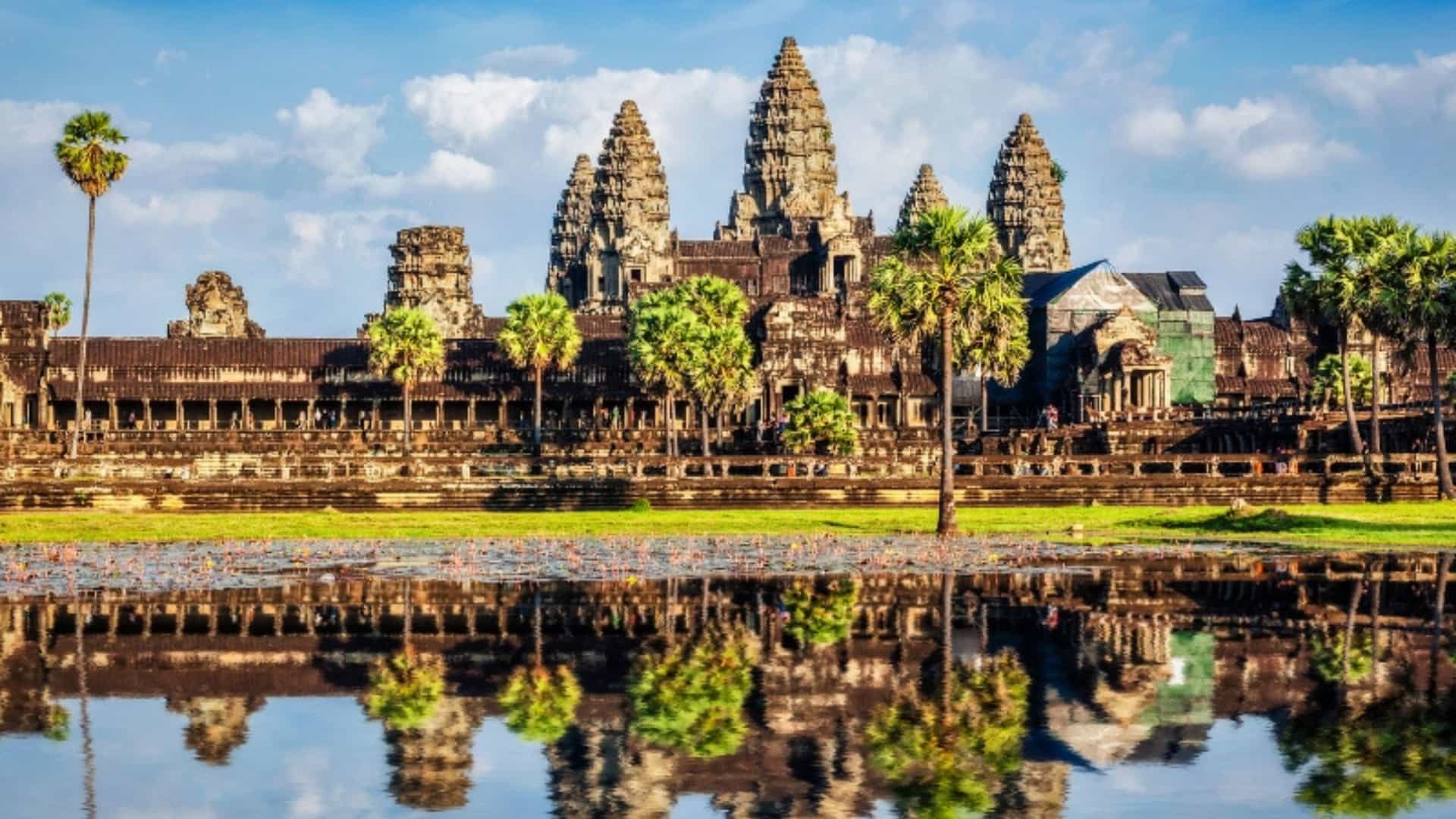 An iconic view of the ancient temple of Angkor Wat in Cambodia, a key landmark in the Southeast Asian river region. The temple’s towering spires and intricate carvings are reflected perfectly in the still water of a large pond in the foreground. Palm trees and lush green grass frame the temple. The sky is a brilliant blue with fluffy white clouds, and the sun shines brightly, highlighting the grand scale and historical significance of the site.