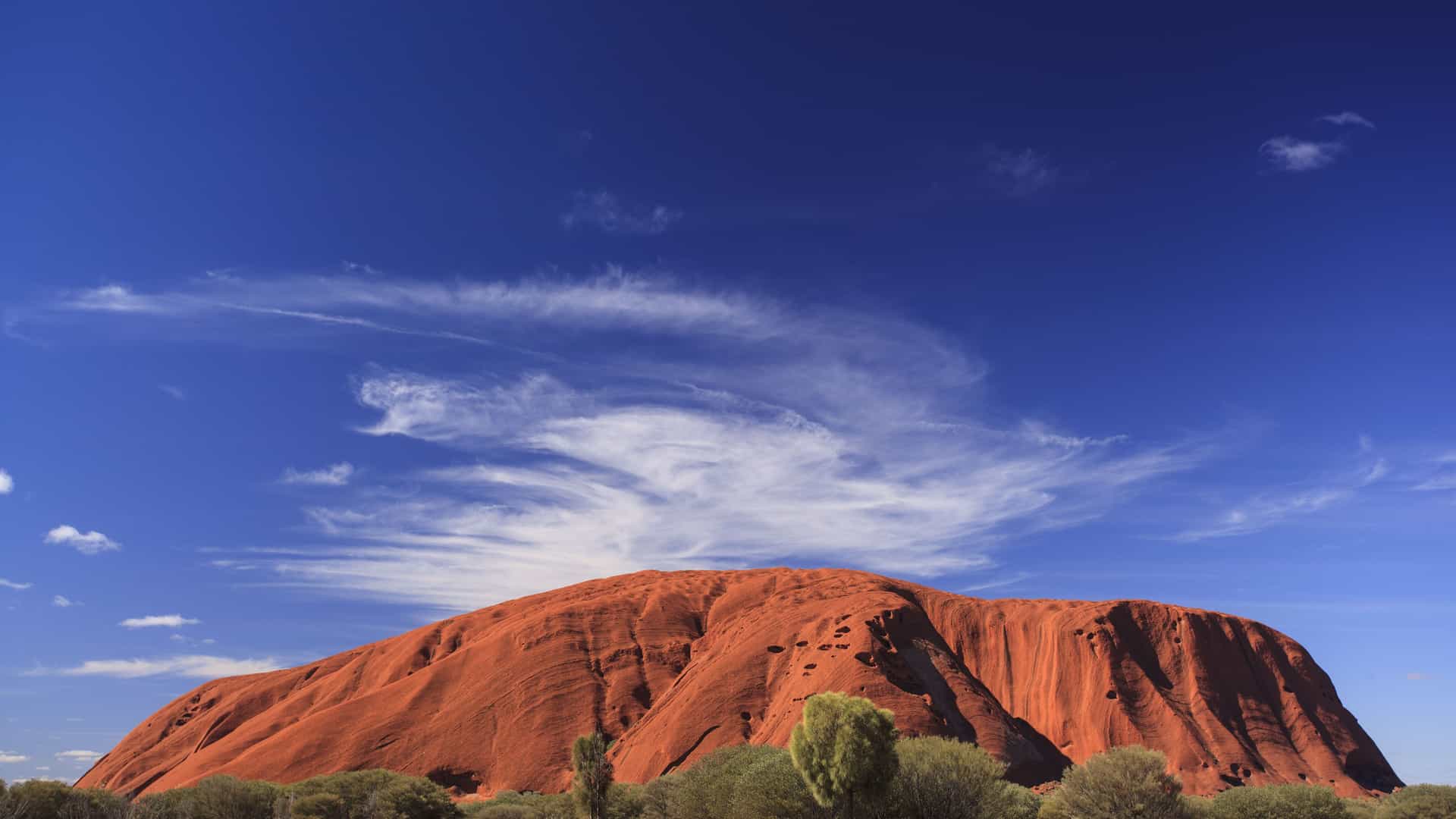 An iconic wide shot of Uluru, also known as Ayers Rock, a massive red sandstone monolith in the heart of Australia's desert landscape. The rock's deep reddish-orange color is prominent against a backdrop of a clear, deep blue sky with wispy white clouds. The foreground is a vast, flat plain covered in dry, golden grass and green scrub bushes. The image captures the immense scale and natural beauty of this sacred landmark in the Australia and New Zealand region.