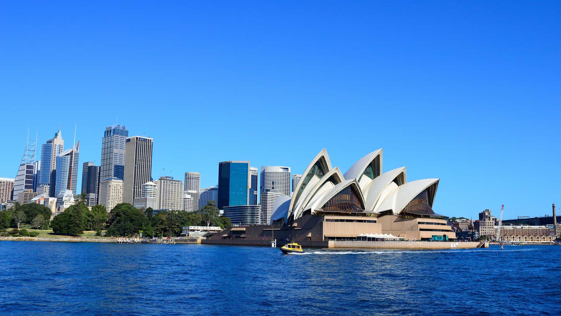 The famous Sydney Opera House stands prominently on the harbor in Sydney, Australia. Its iconic white, shell-like sails are a striking contrast against the clear blue sky. In the background, the modern skyline of downtown Sydney, with its collection of glass and steel skyscrapers, rises from the shore. The dark blue water of the harbor is calm, with a small yellow boat passing in the foreground. The image captures a quintessential view of this major urban landmark in the Australia and New Zealand region.