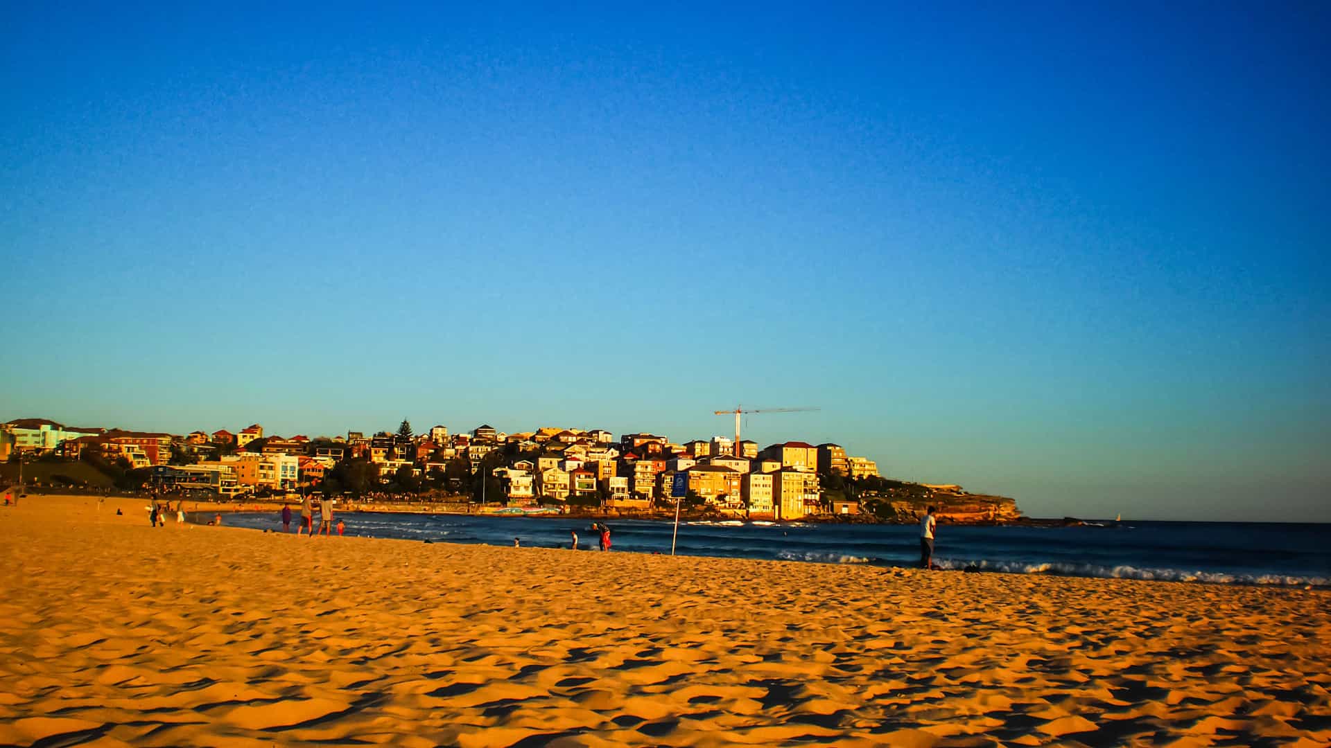The vast, golden sand of Bondi Beach in Sydney, Australia, is illuminated by the late afternoon sun. The sand is textured with ripples and footprints leading toward the gentle waves of the blue ocean. In the background, a cluster of buildings, including hotels and apartments, sits on a hill overlooking the beach. The sky is a gradient of deep blue to a lighter shade on the horizon. The image captures the serene and iconic coastal landscape of the Australia and New Zealand region.