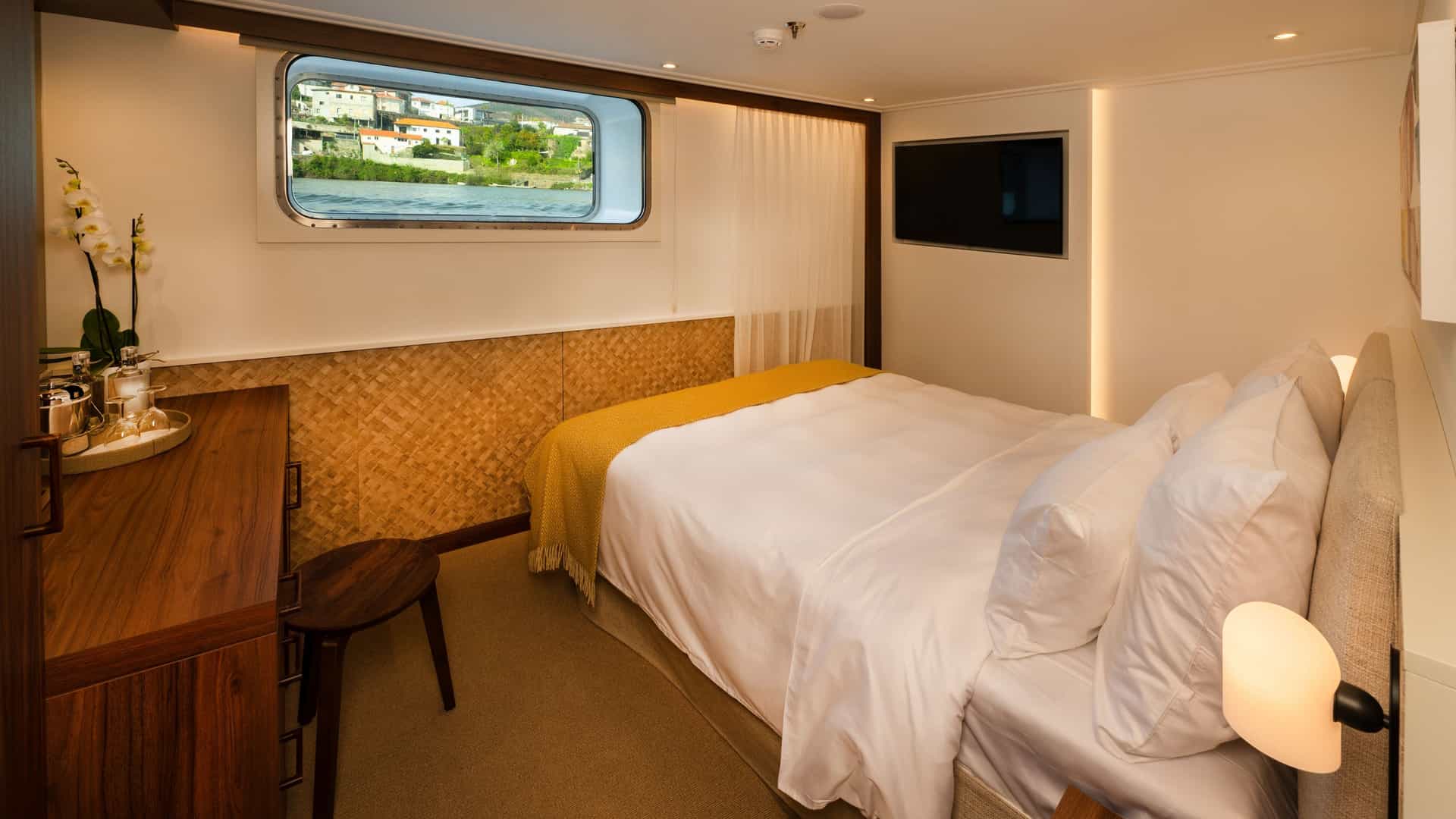 Deluxe stateroom on an Avalon Waterways river cruise ship featuring a double bed, large panoramic window overlooking a scenic riverbank town, wooden vanity desk, and wall-mounted flat-screen TV.