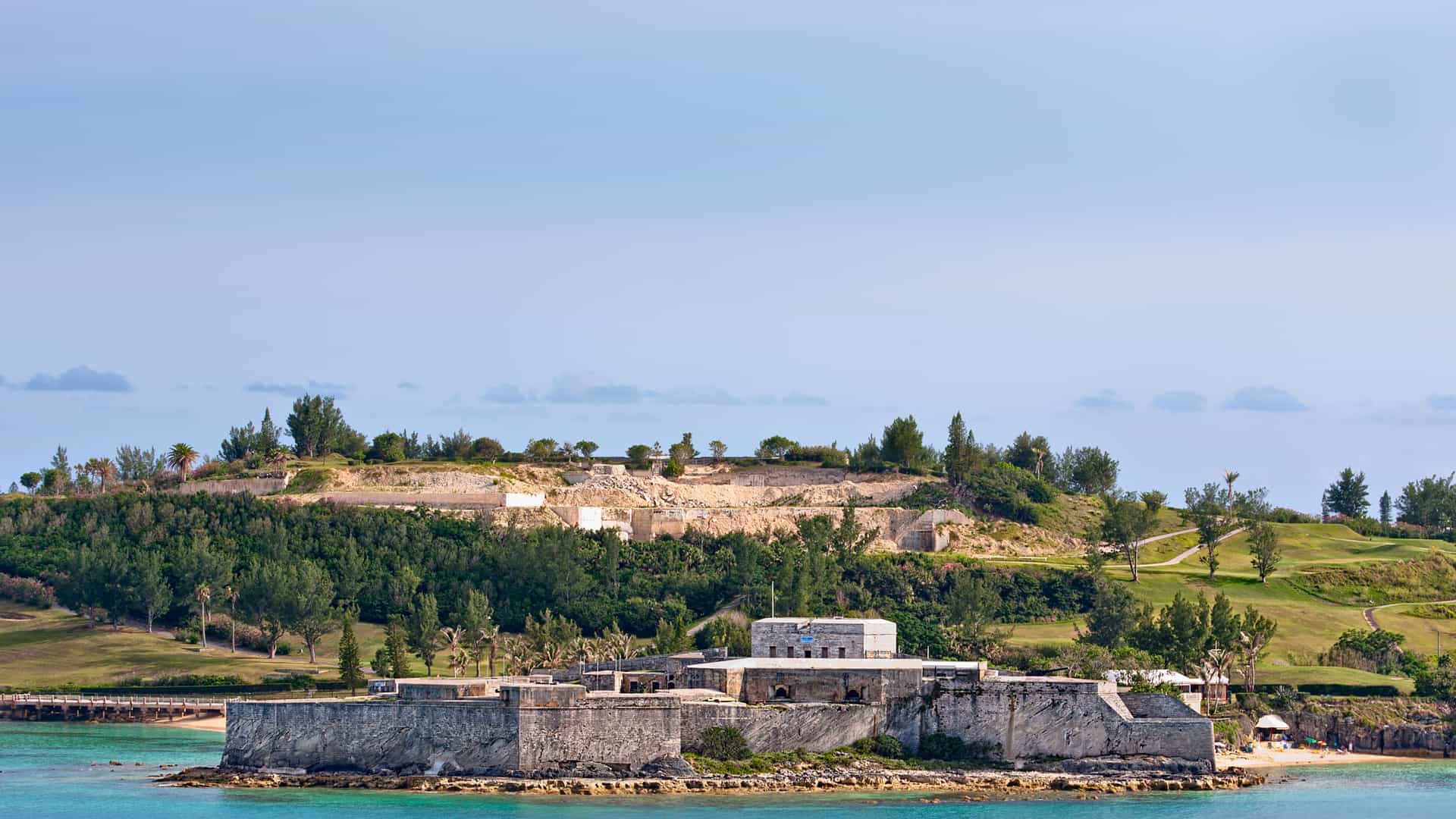Fort St. Catherine, a historic fortress on St. George's Island in Bermuda, is seen from the water. The fort's gray stone walls and blockhouse are set on a small peninsula surrounded by clear, turquoise water. The building has a flat roof and is surrounded by a low stone wall. A lush green hill rises behind the fort, covered in trees and some open, grassy areas. The sky is a pale blue with a few scattered clouds, highlighting the historical architecture and scenic beauty of this Bermuda landmark.