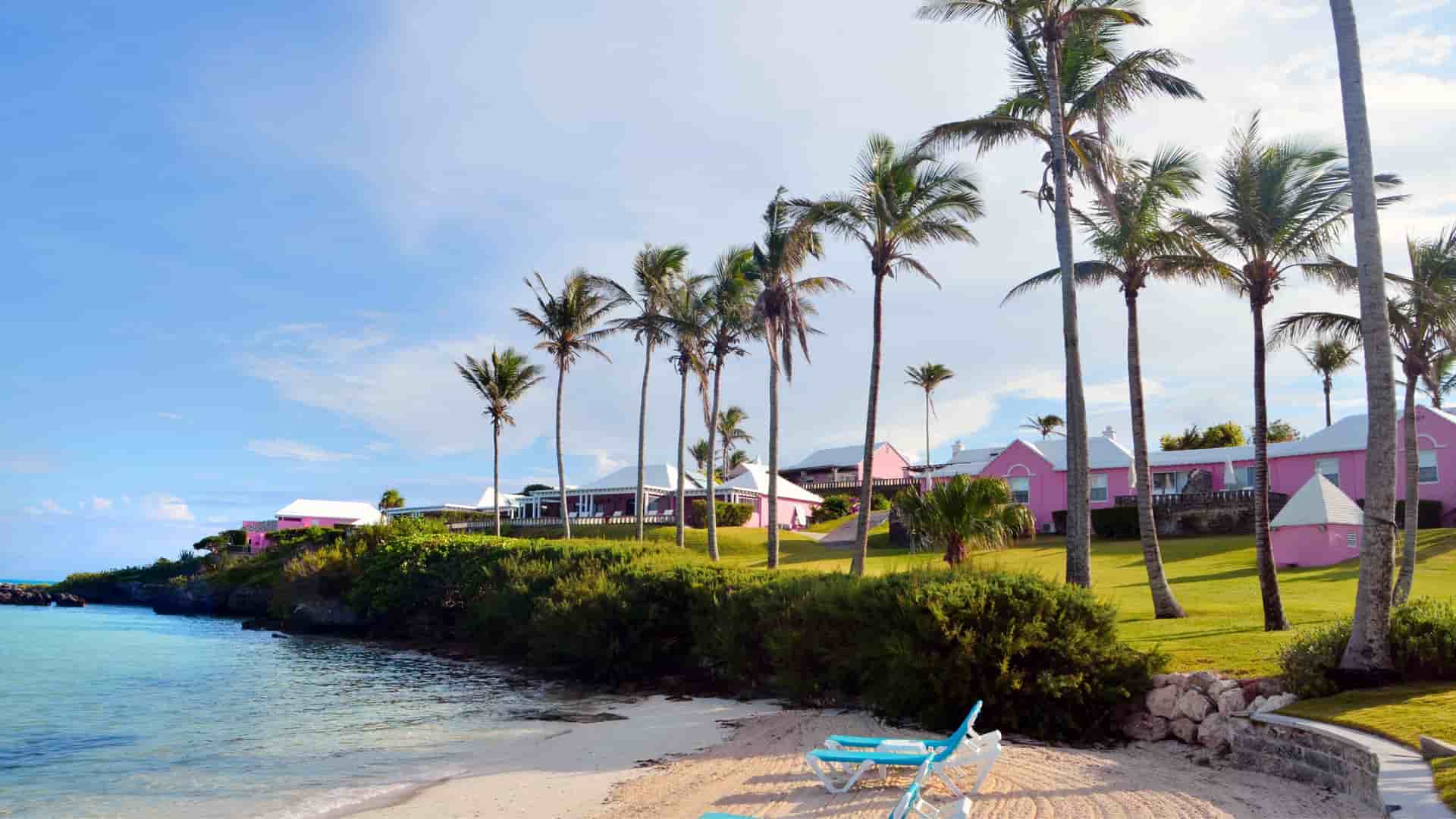 Teal colored lounge chairs on the beach with palm trees and pink beach cottages in the background.
