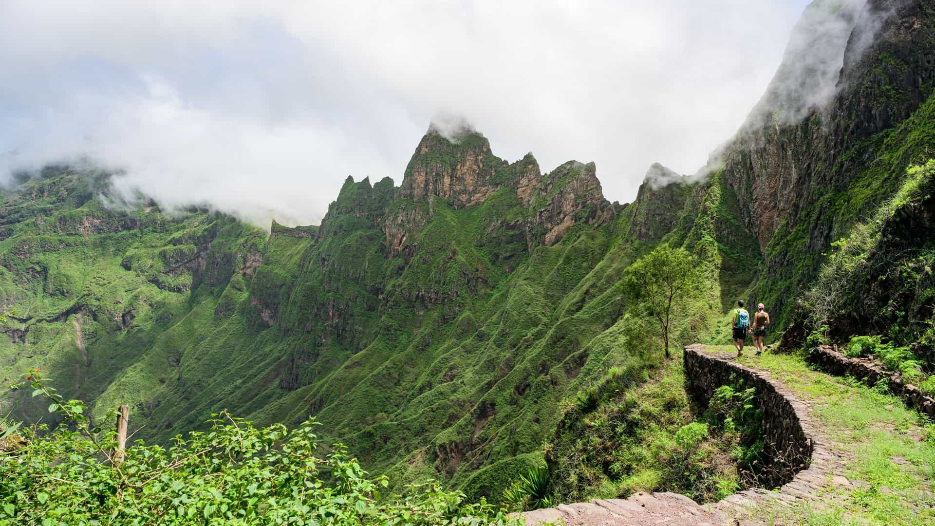 Two hikers walk along a winding stone path overlooking the lush, dramatic green volcanic ridges of the Paúl Valley in Santo Antão, Cape Verde, under a mist-shrouded sky.