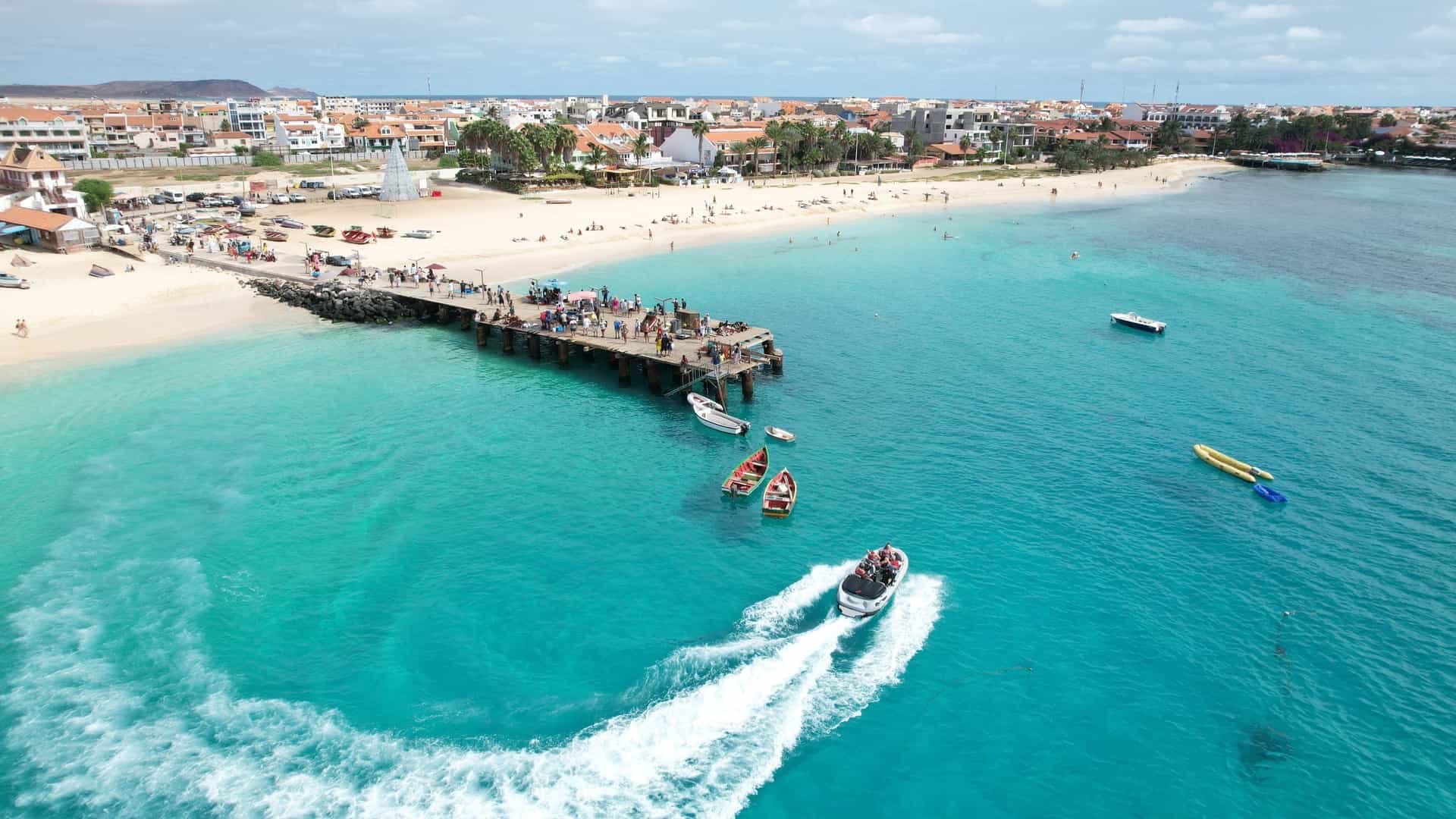 An aerial, high-angle view captures a speedboat leaving a white wake in vibrant, turquoise ocean water near a wooden pier crowded with people alongside a white-sand beach and the coastal town of Santa Maria.