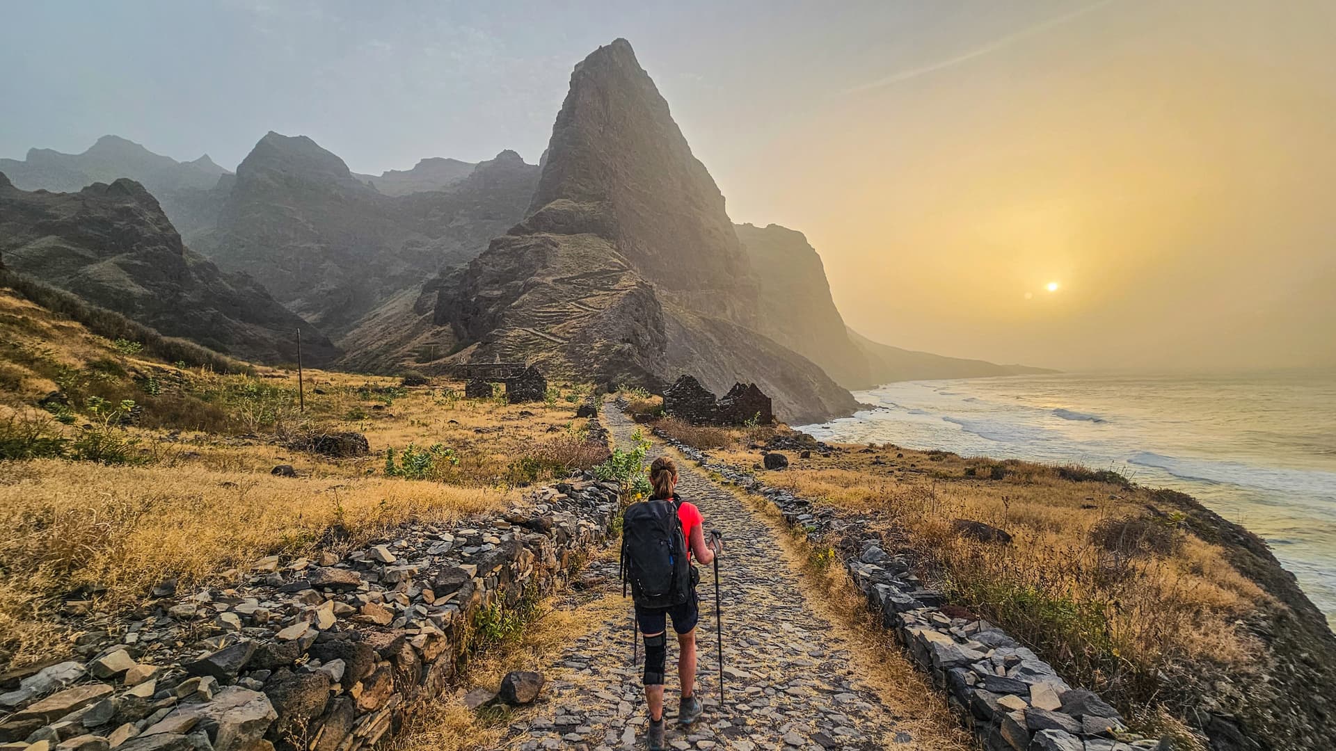 A hiker with a backpack and trekking poles walks along a historic stone-cobbled coastal path at sunset, with dramatic volcanic mountains on the left and the Atlantic ocean waves crashing on the right.