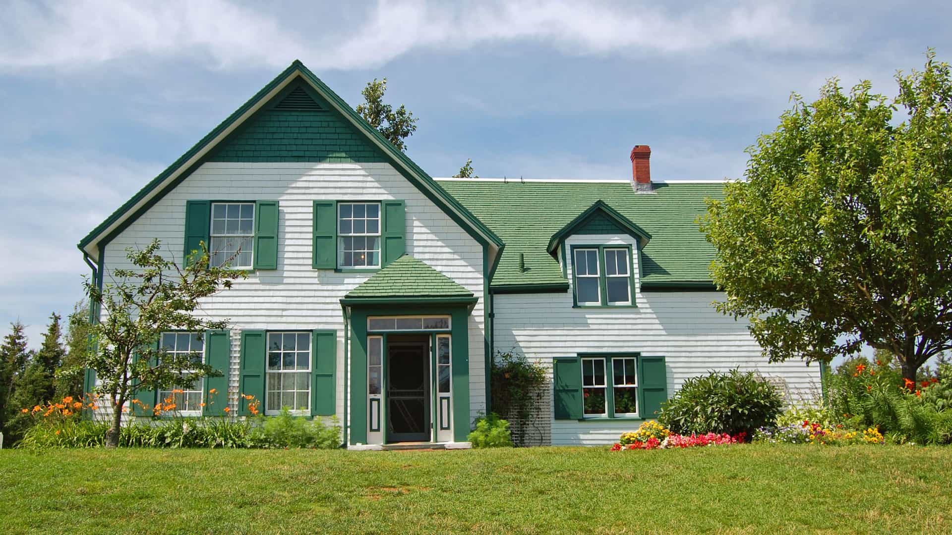 The iconic Green Gables house on Prince Edward Island, Canada, the setting of the 'Anne of Green Gables' novels. The two-story white farmhouse features a distinctive green roof and green shutters. The front yard is a lush green lawn, with a few trees and bushes adorned with colorful flowers. The sky above is bright blue with scattered white clouds, creating a peaceful and classic scene that represents a key cultural landmark of the Canada and New England region.