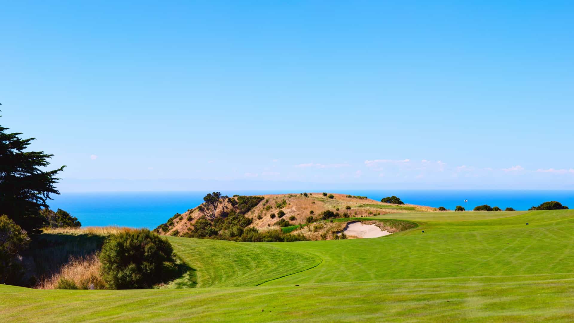 Sunlight shining on the rolling green fairways and a deep sand bunker at Cape Kidnappers golf course in New Zealand, set against the bright blue Pacific Ocean.