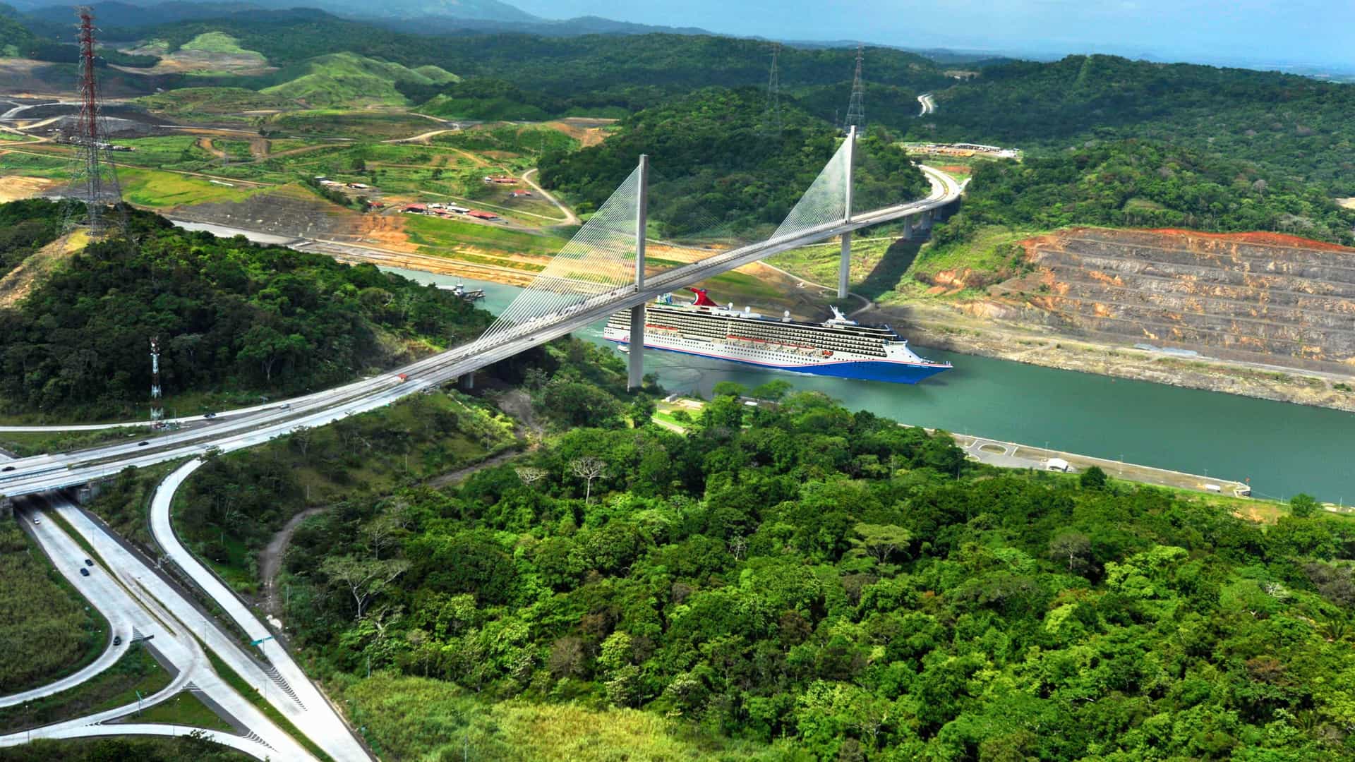 Carnival Spirit-class cruise ship passing under a modern bridge in the Panama Canal, surrounded by lush landscapes.
