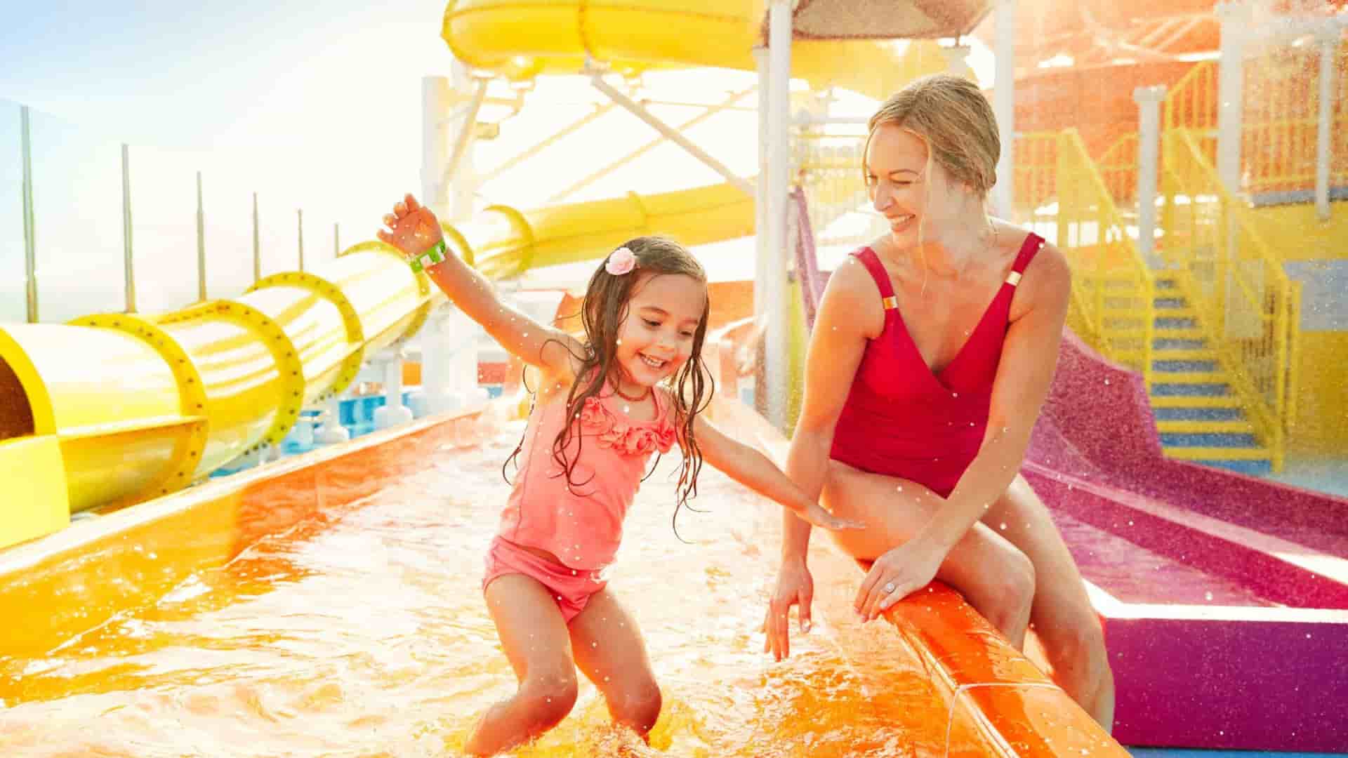 A mother and daughter laughing while playing in a vibrant water park with yellow and orange slides on a Carnival Cruise ship