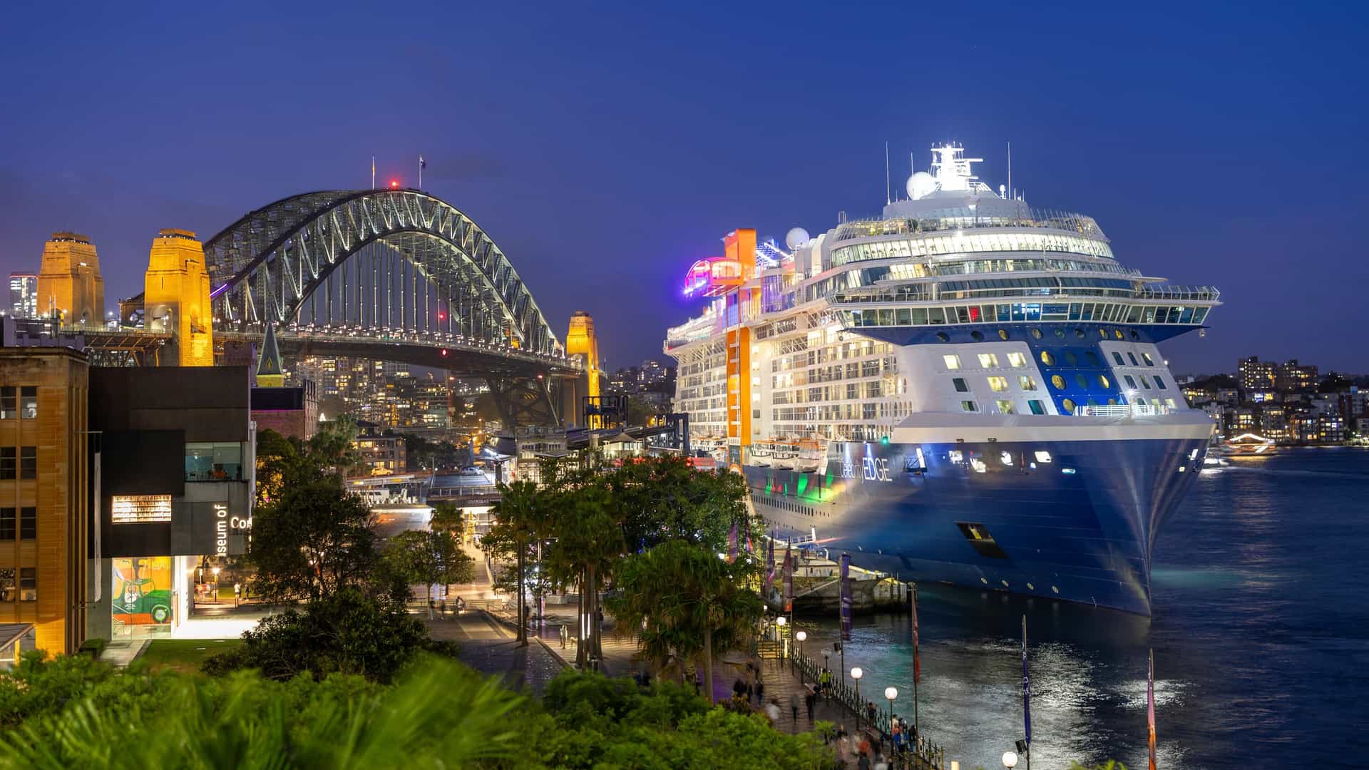 Celebrity Edge cruise ship lit up overnight at Sydney's Circular Quay, with the Sydney Harbour Bridge glowing behind it against a deep blue evening sky.