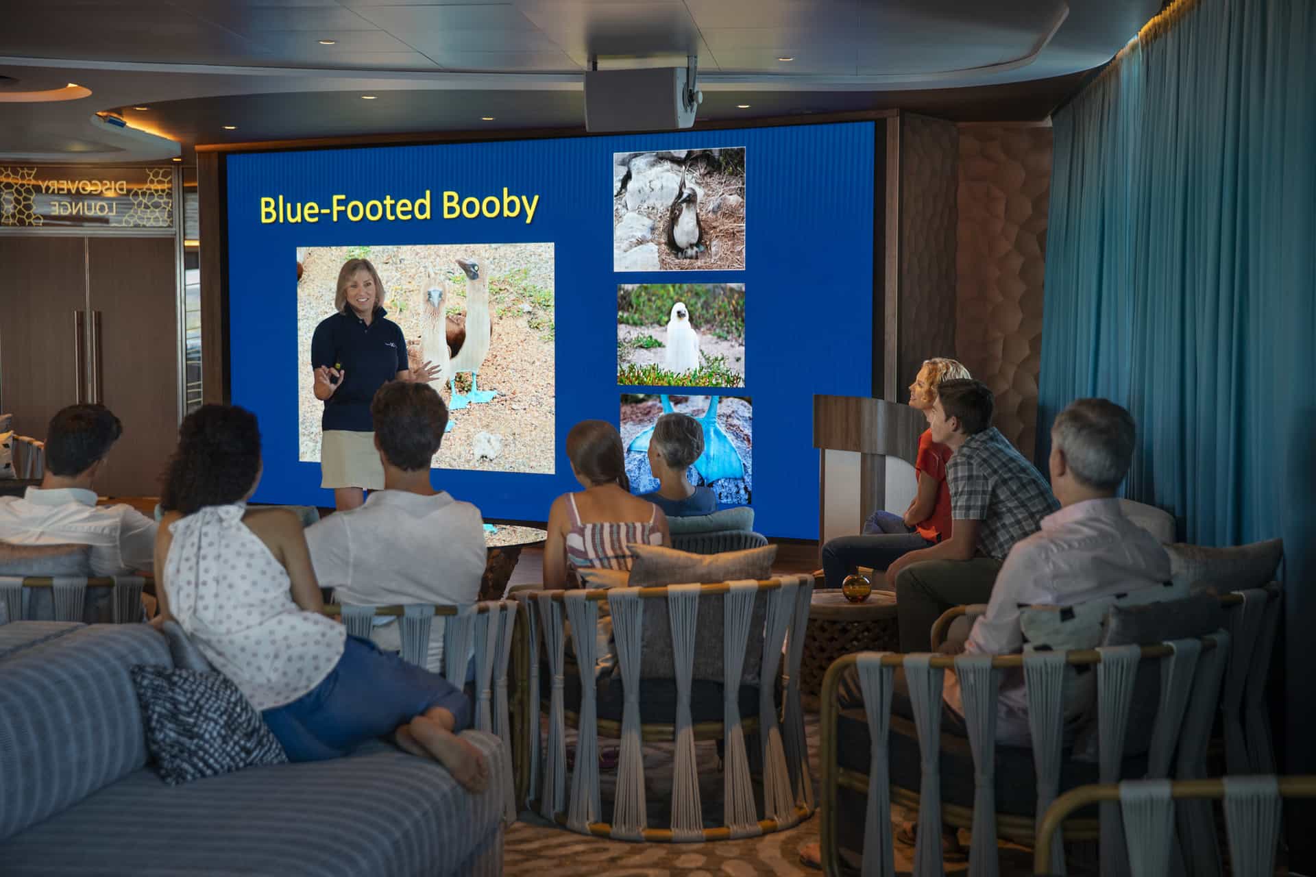 Guests relax in the modern, earth-toned Discovery Lounge of the Celebrity Flora, where a naturalist stands at a podium in front of a large digital screen presenting a map of the Galapagos Islands.