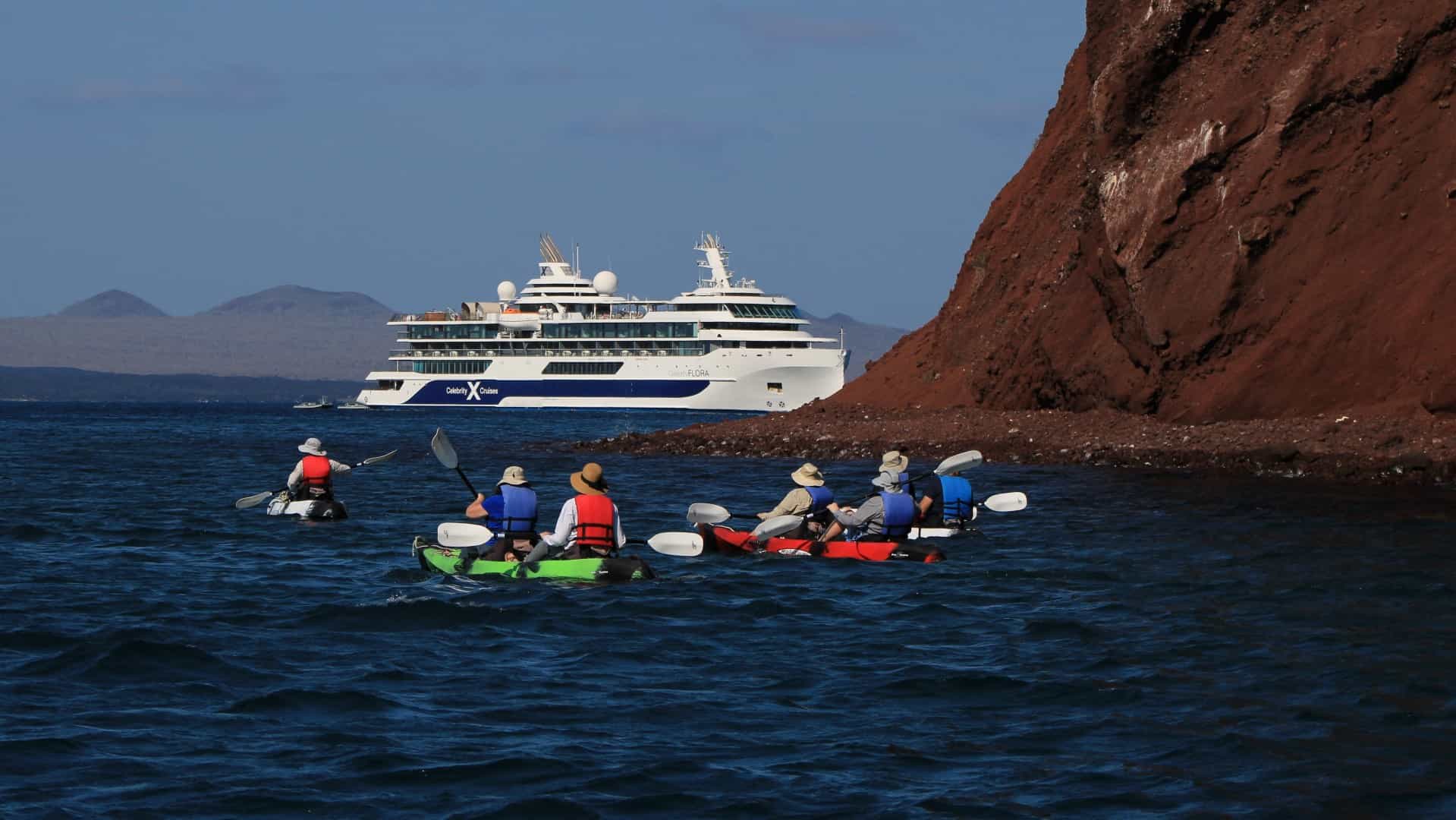 Several colorful kayaks with passengers paddle across deep blue water toward the towering, rusty-red volcanic cliffs of Rabida Island, with the white Celebrity Flora mega-yacht anchored in the background.