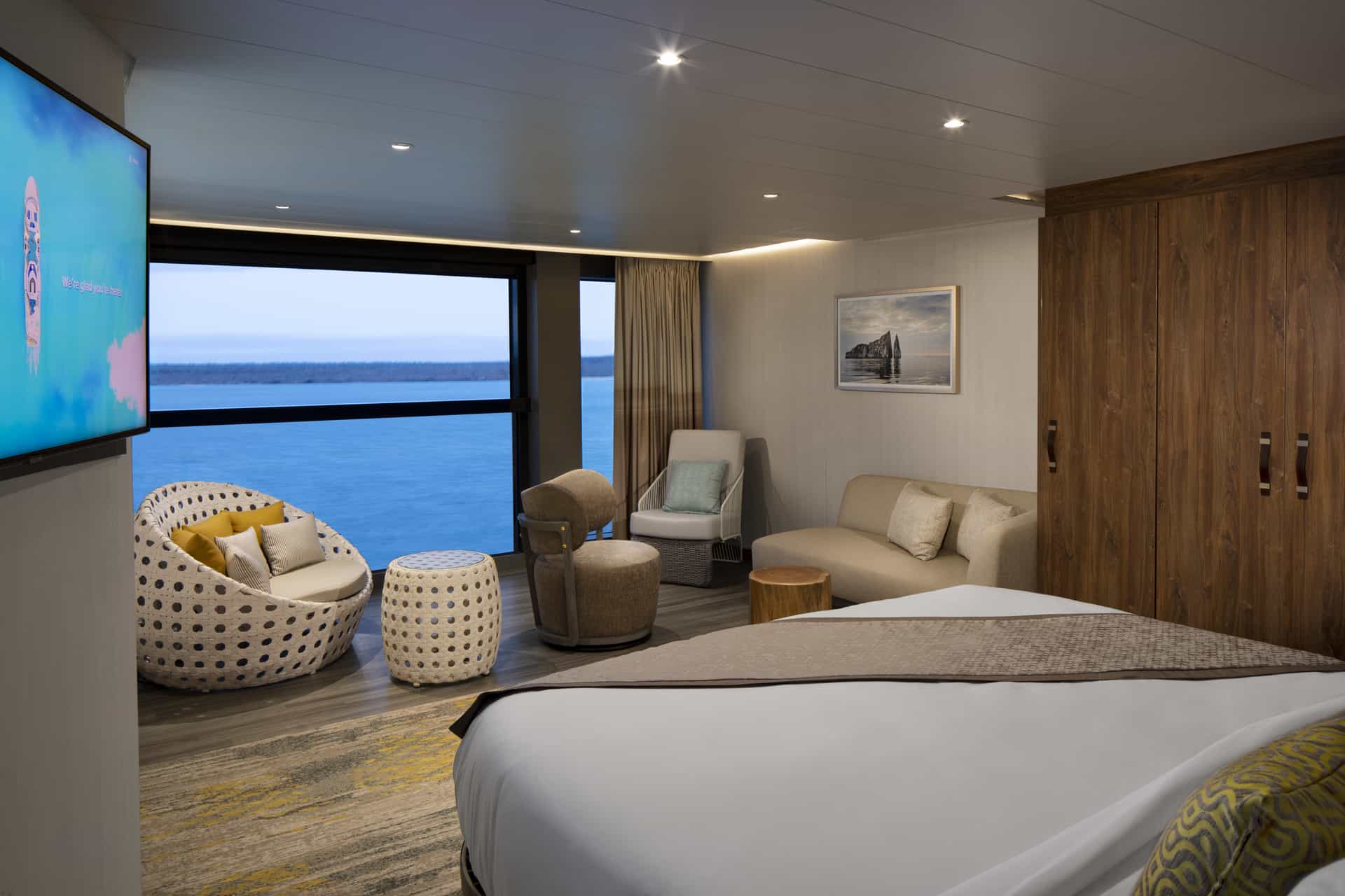 A luxurious, modern suite, aboard Celebrity Flora, features organic-style furniture and a large bed facing an "Infinite Veranda" window that offers a panoramic view of the ocean at twilight.