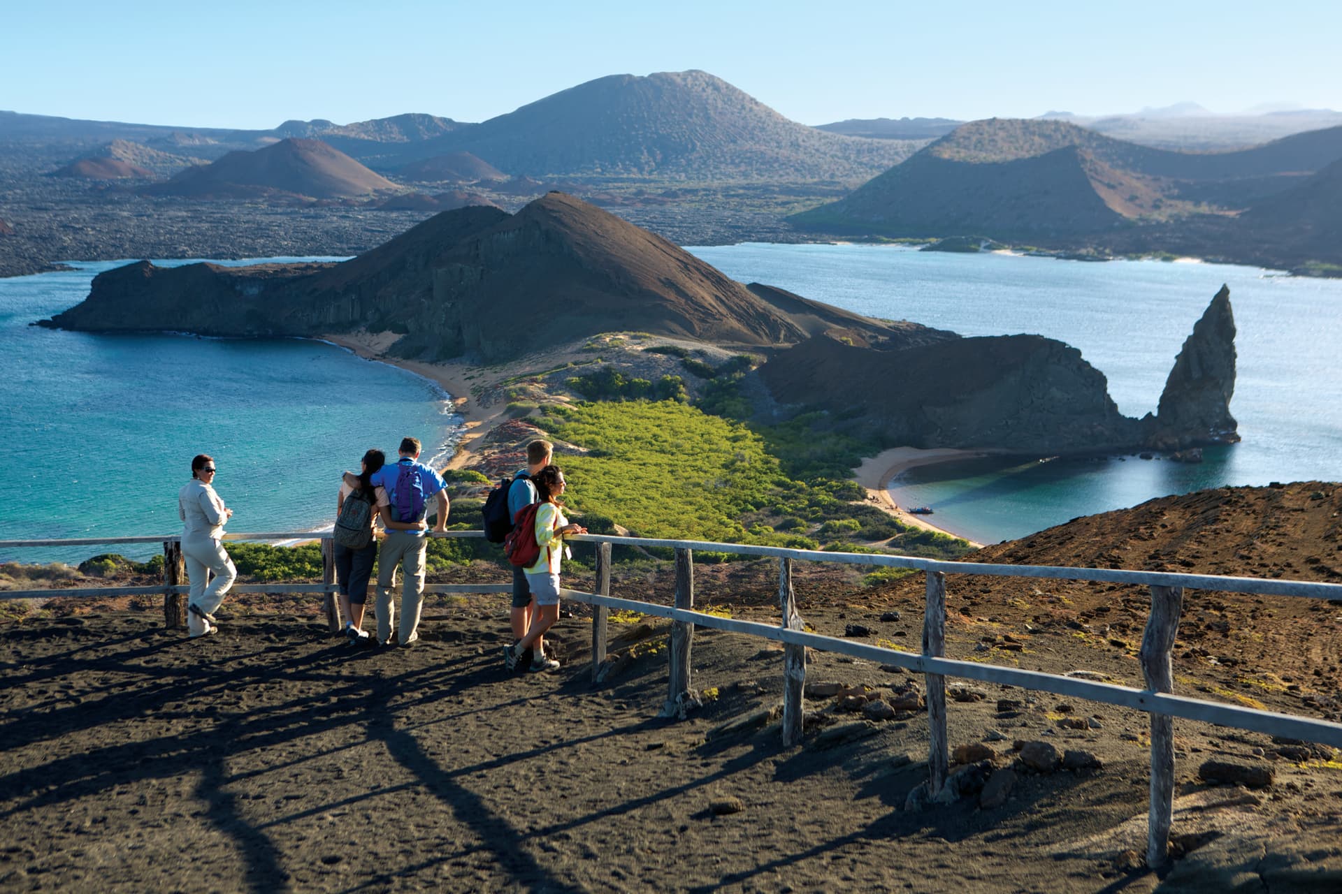 A group of five travelers stands behind a rustic wooden railing atop a black volcanic hill, gazing across a mangrove-lined isthmus toward the iconic, sharp-pointed Pinnacle Rock rising from turquoise waters amidst ancient craters.