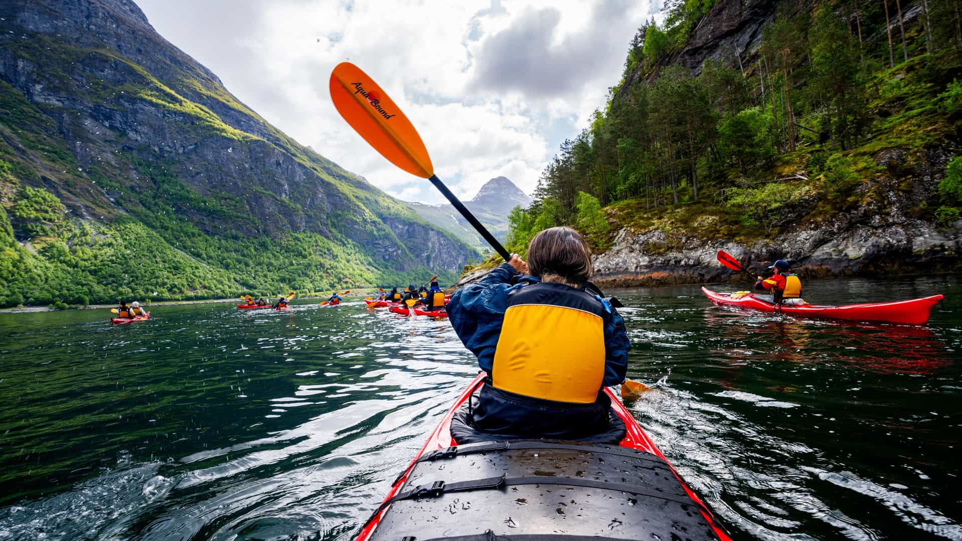 Kayakers paddling through the calm waters of Geirangerfjord, Norway, surrounded by towering cliffs and lush green mountainsides.