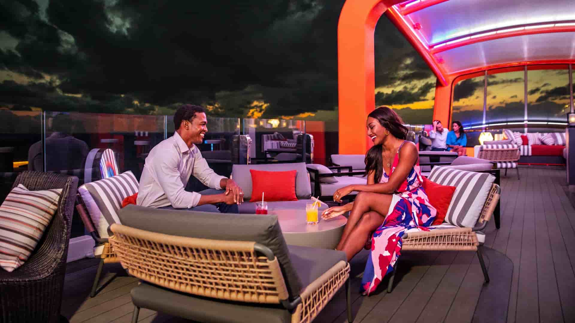 A couple on the Celebrity Cruises' Celebrity Xcel ship is sitting on a modern outdoor lounge deck with a bright orange bar overhead, laughing and facing each other over drinks while the sun sets behind them.