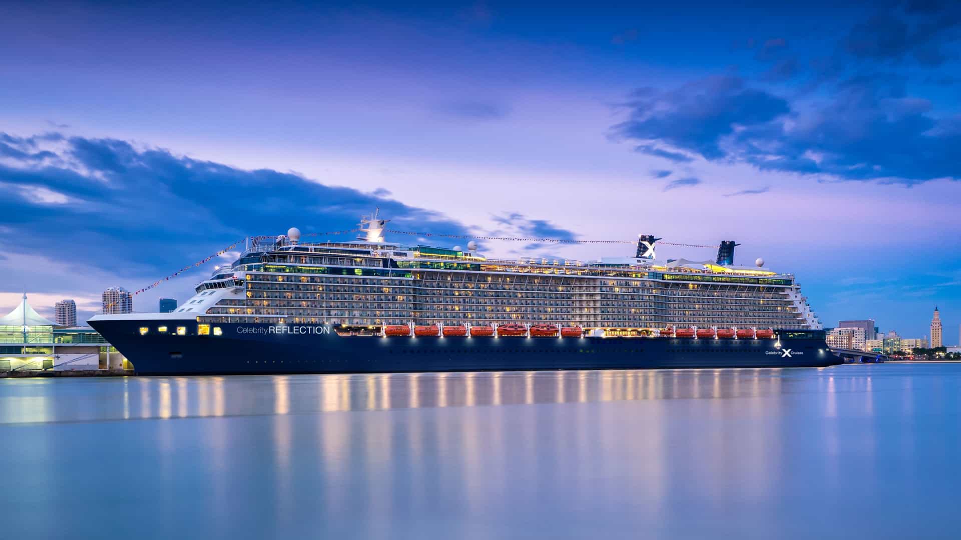 Exterior side profile of the Celebrity Reflection cruise ship docked at a port in Miami during twilight with its lights glowing and reflecting on calm water.