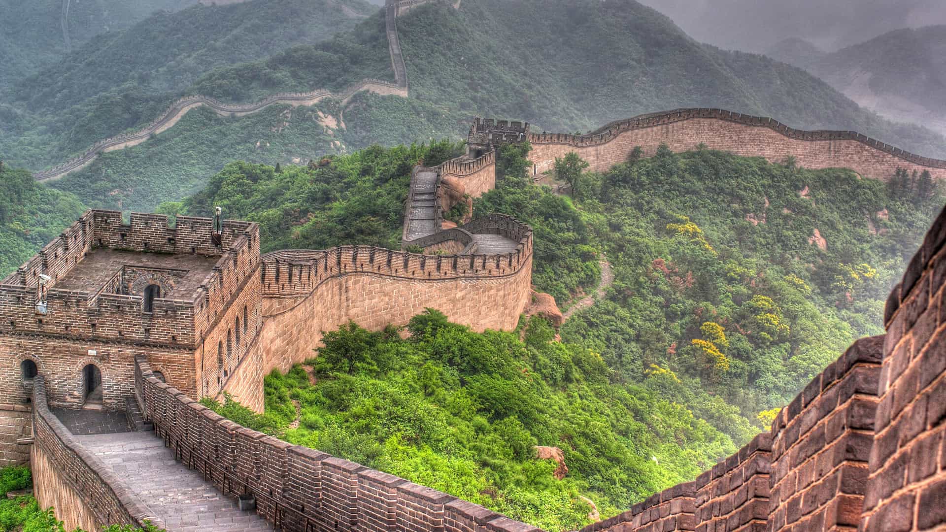 An atmospheric view of the Great Wall of China as it winds its way over lush, green, fog-covered mountains. The stone and brick wall, with its numerous watchtowers, snakes up and down the steep terrain, disappearing into the mist in the distance. The mountains are covered in thick trees and foliage, with rocky outcrops visible through the greenery. The image captures the impressive scale and historical grandeur of this famous landmark in a dramatic, foggy Chinese landscape.