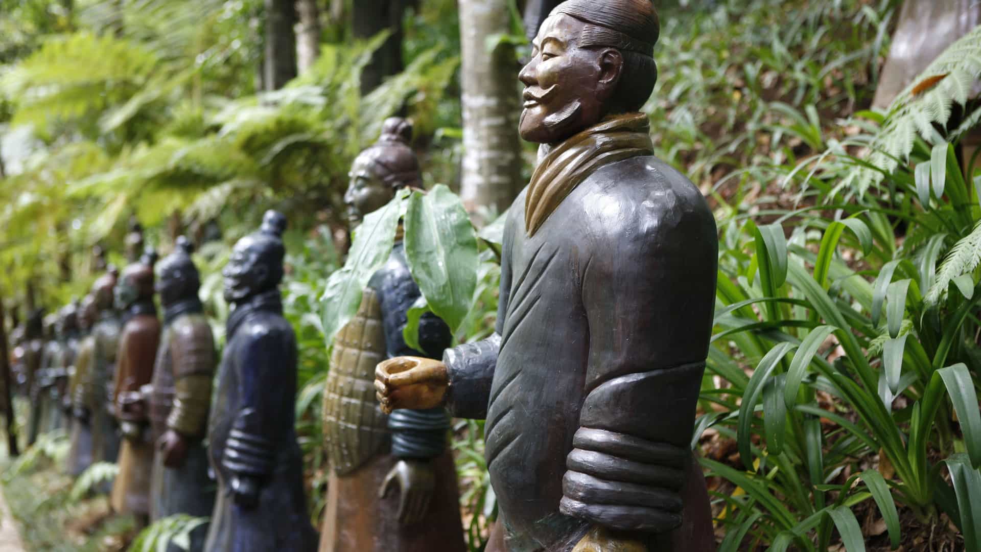 A row of small replicas of the famous Terracotta Warriors is set in a lush garden. The statues are detailed, showing the varied faces and armor of the original figures. They are lined up on a path, surrounded by vibrant green ferns and other tropical plants. The scene, which is likely in Xi'an, China, depicts the popular attraction where the famous archaeological find is on display, representing the rich history of the China region.