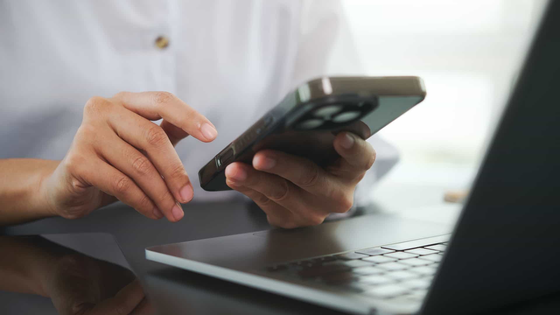A close up shot of hands using a smartphone and laptop at a desk.