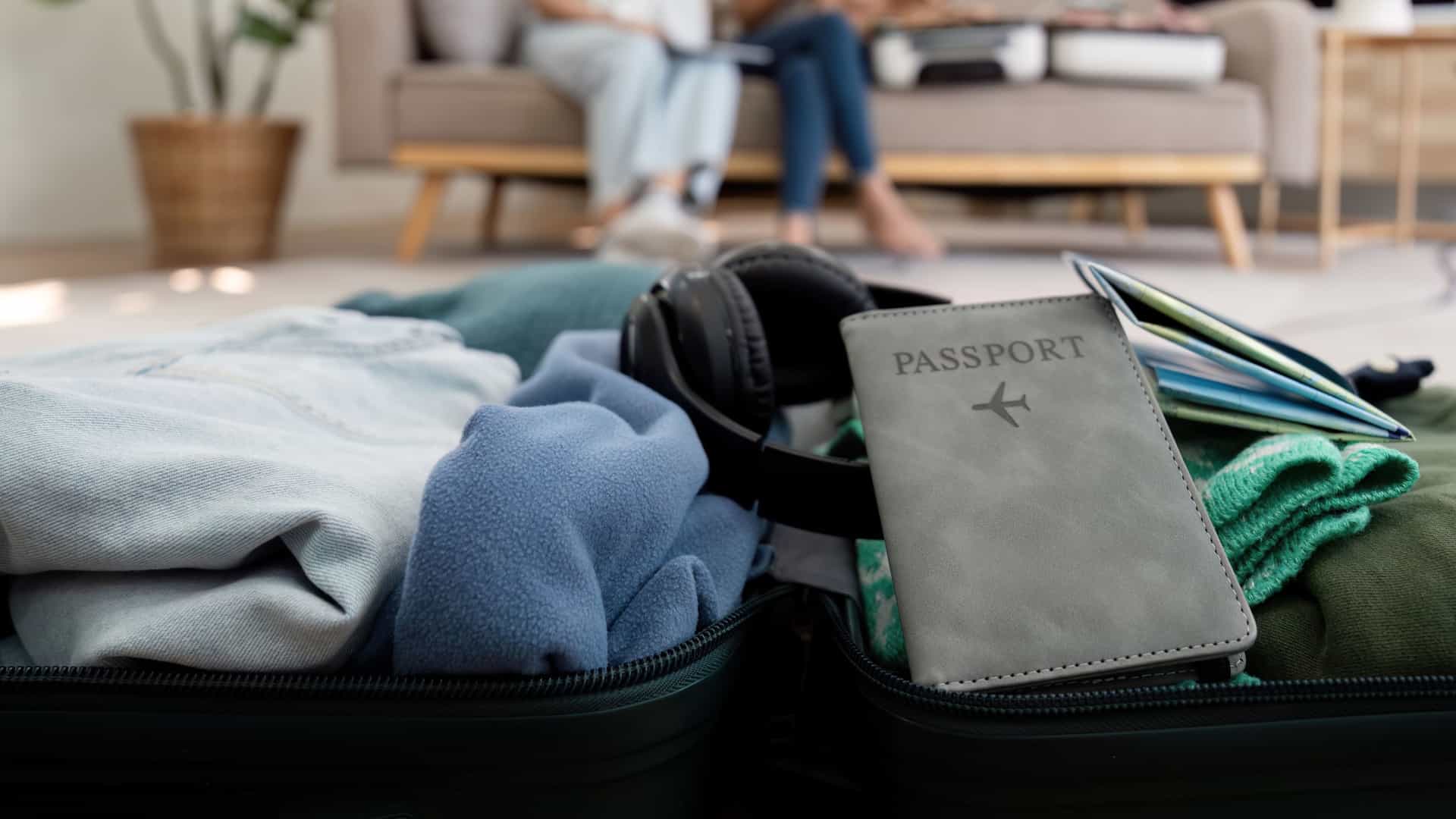 Close up shot of a suitcase with passport, headphones, and clothes inside with a two people sitting on the couch in the background.