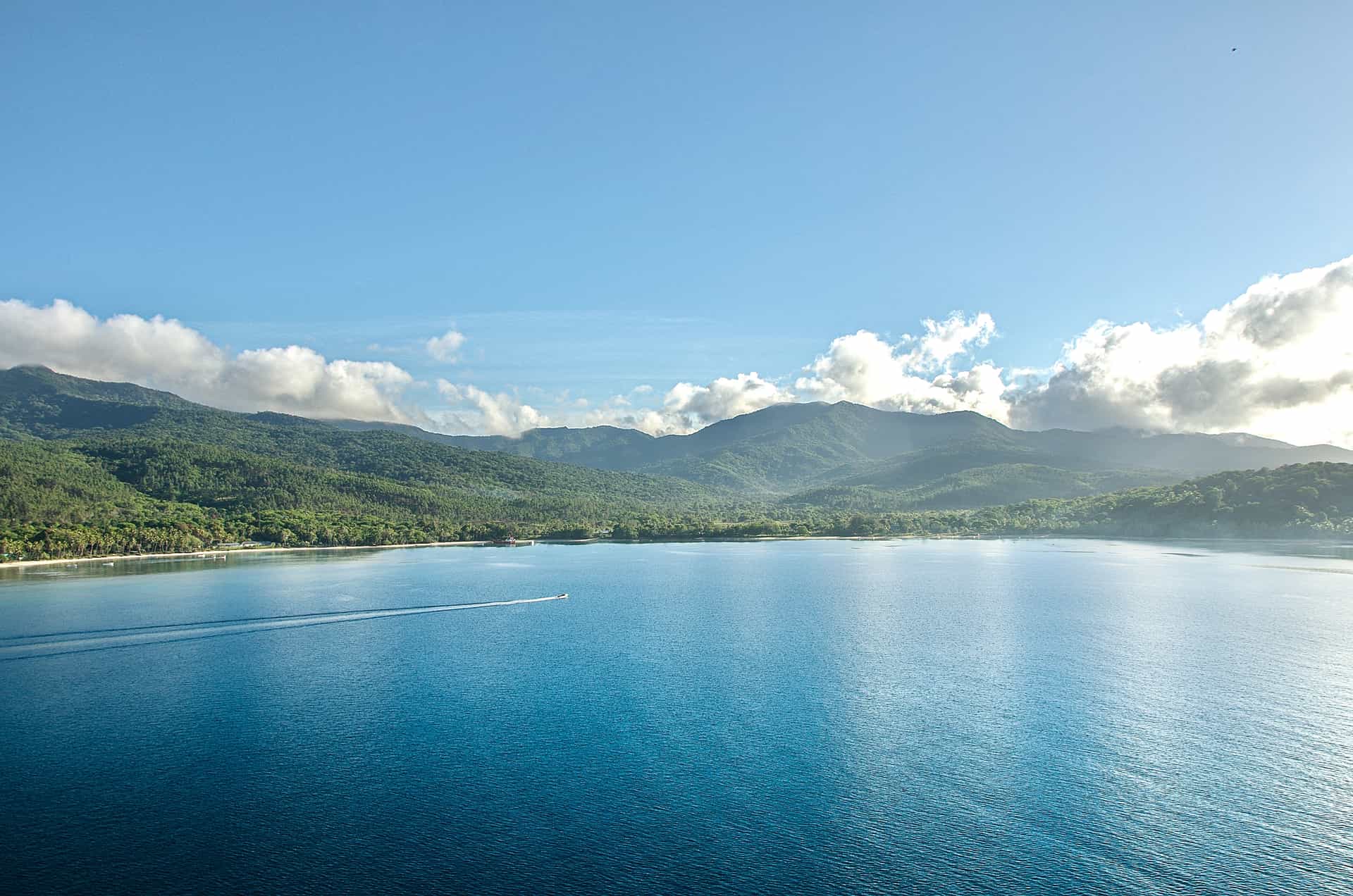 An aerial view of the serene blue waters of Anelghowhat on Aneityum Island, Vanuatu, a lush South Pacific cruise port destination featuring dense green tropical mountains under a bright, partly cloudy sky.