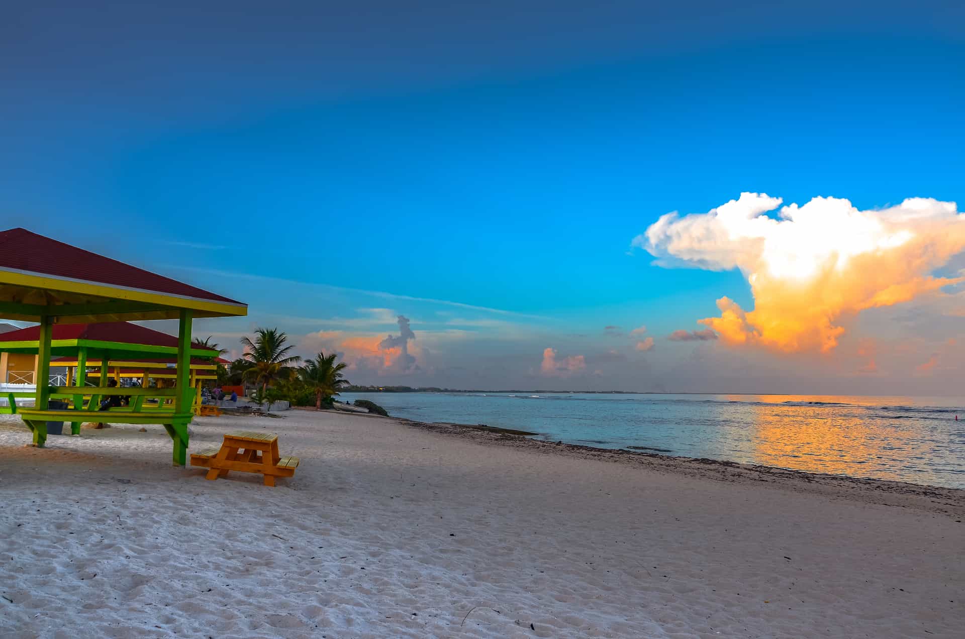 A scenic view of the sandy shore at Bodden Town, a popular cruise excursion destination on Grand Cayman in the Cayman Islands, featuring bright green and red beach pavilions alongside a dramatic Caribbean sunset sky reflecting off the ocean.