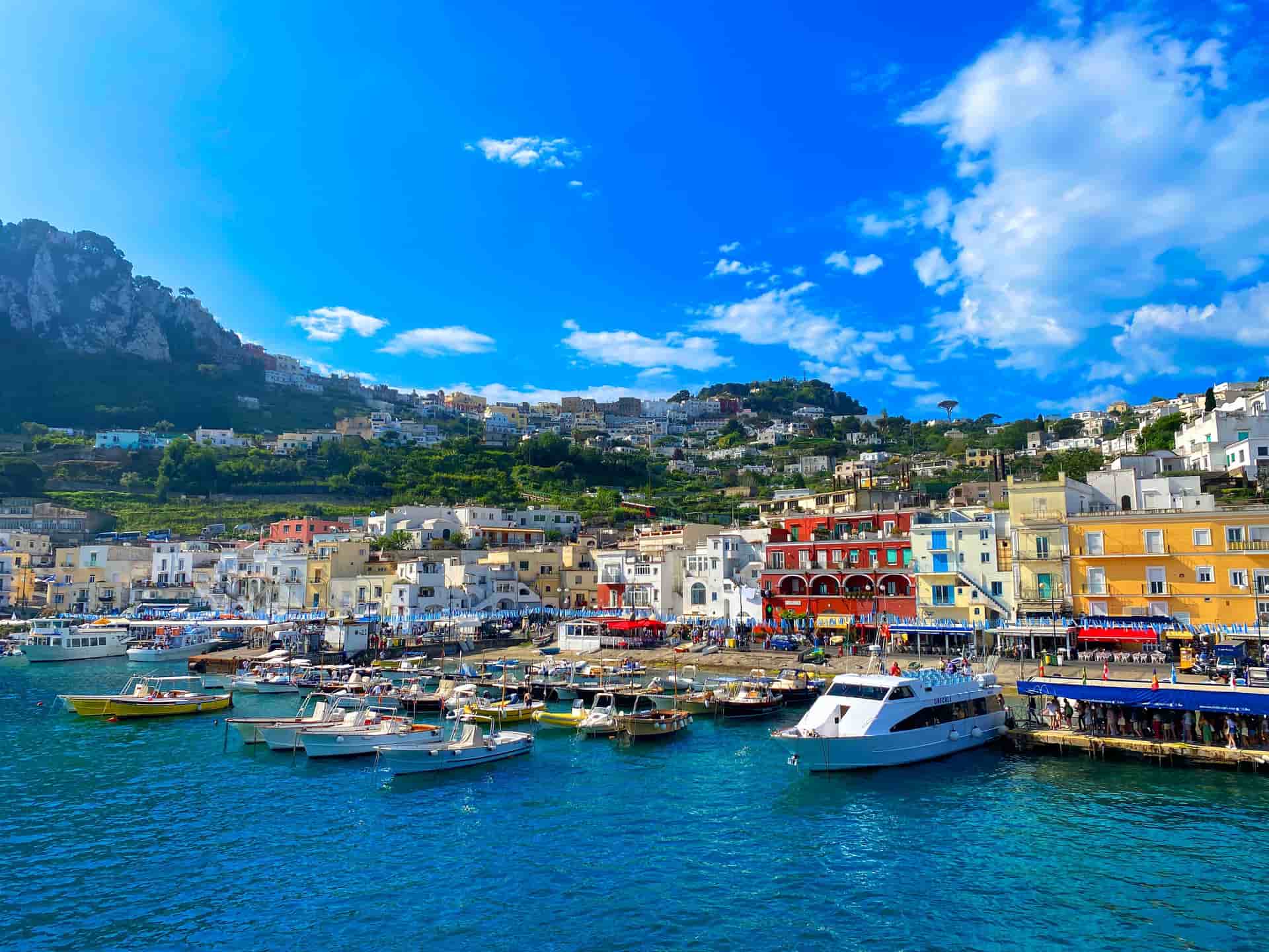A vibrant view of Marina Grande, the bustling harbor on the island of Capri, Italy—a classic shore excursion from the Sorrento and Naples cruise ports. The image shows numerous boats and ferries docked in clear turquoise waters, framed by colorful buildings and lush, steep cliffs rising into a bright blue sky.