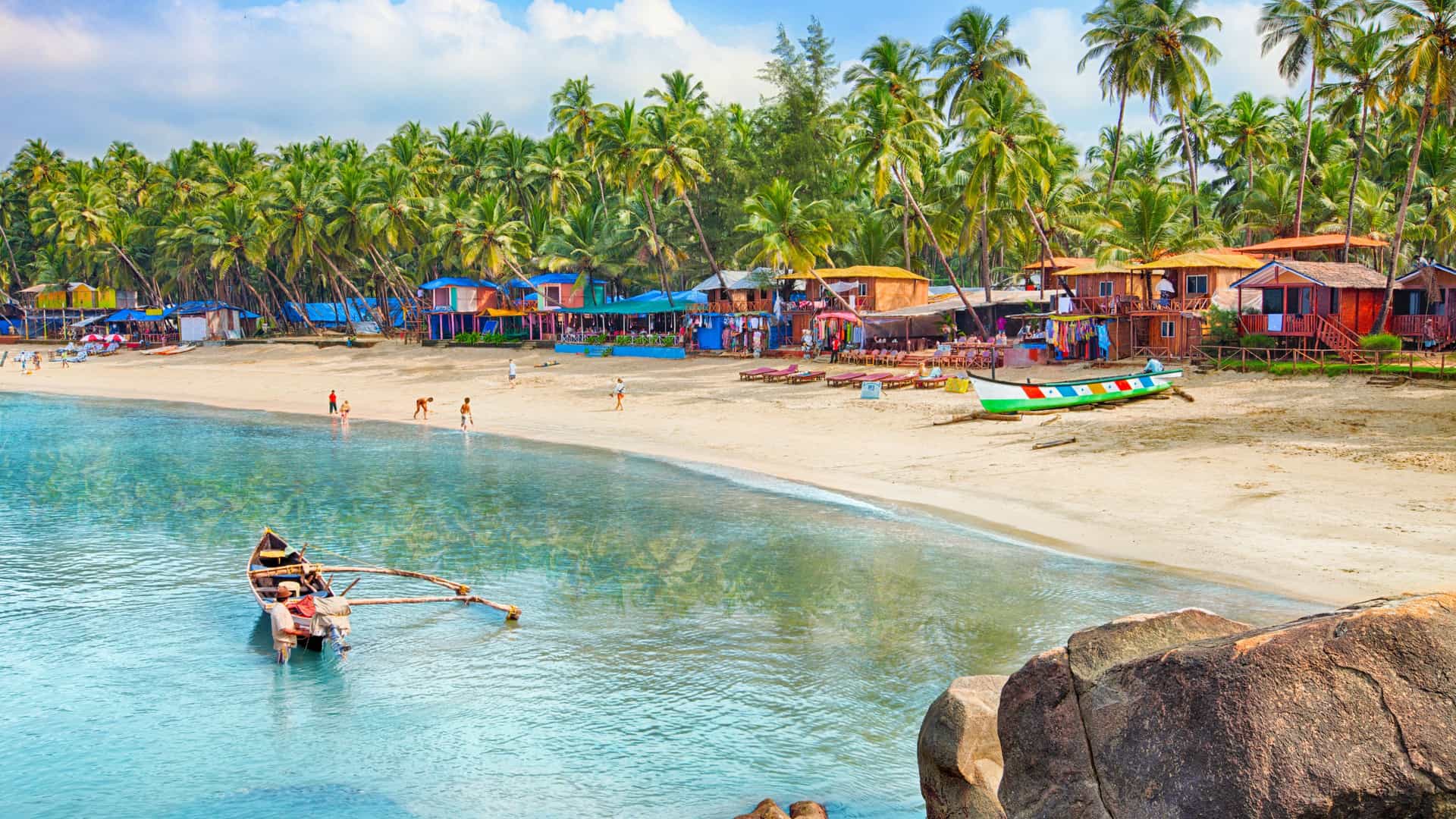 A tropical beach in Goa with turquoise water, palm trees, colorful huts, and wooden boats along a sunny shoreline.