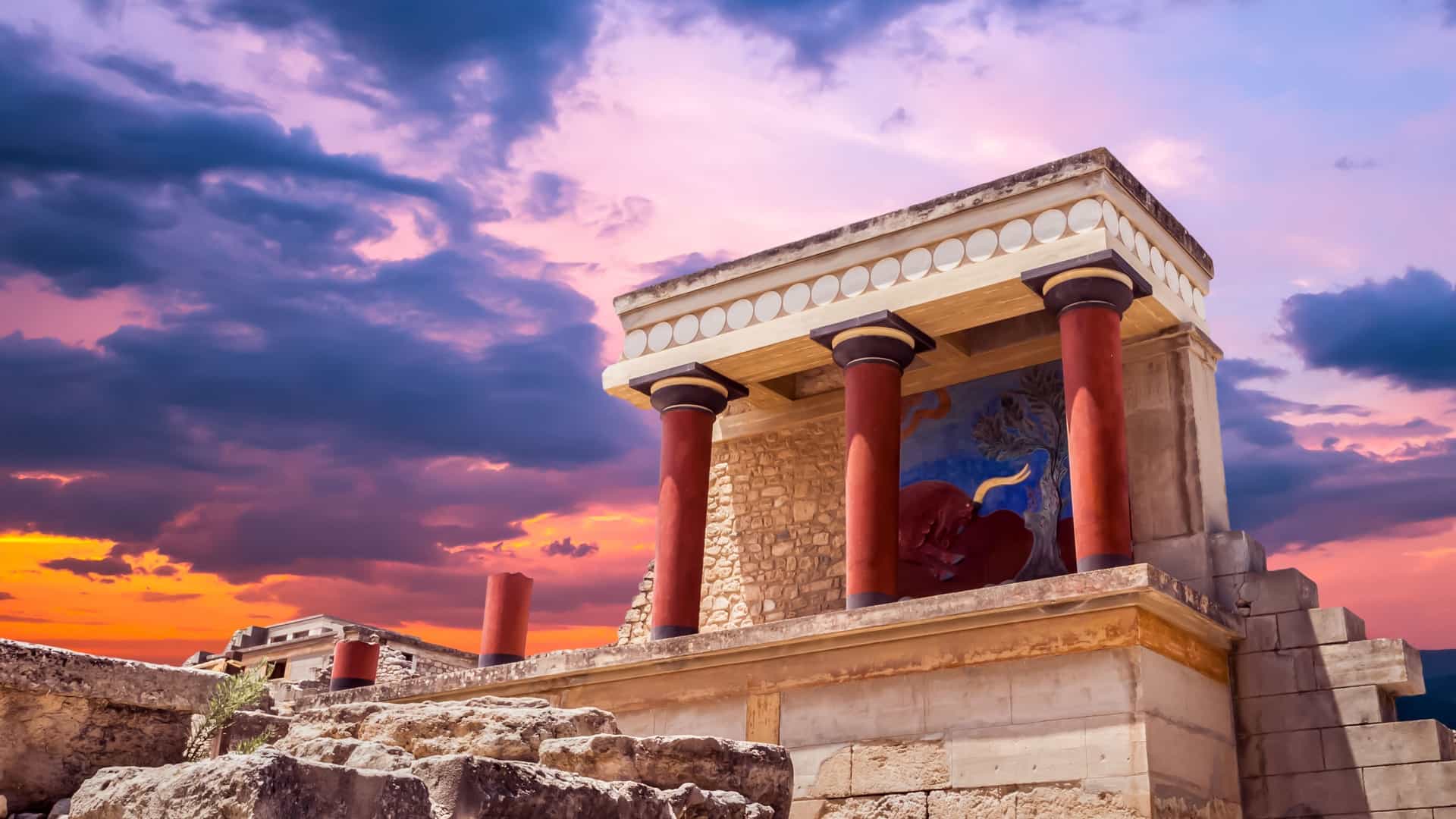 The ancient ruins of the Palace of Knossos, a premier archaeological shore excursion destination near the Heraklion (Iraklion) cruise port on the island of Crete, Greece. The image highlights the reconstructed North Bastion with its iconic red columns and charging bull fresco, set against a dramatic sunset sky filled with purple and orange clouds.