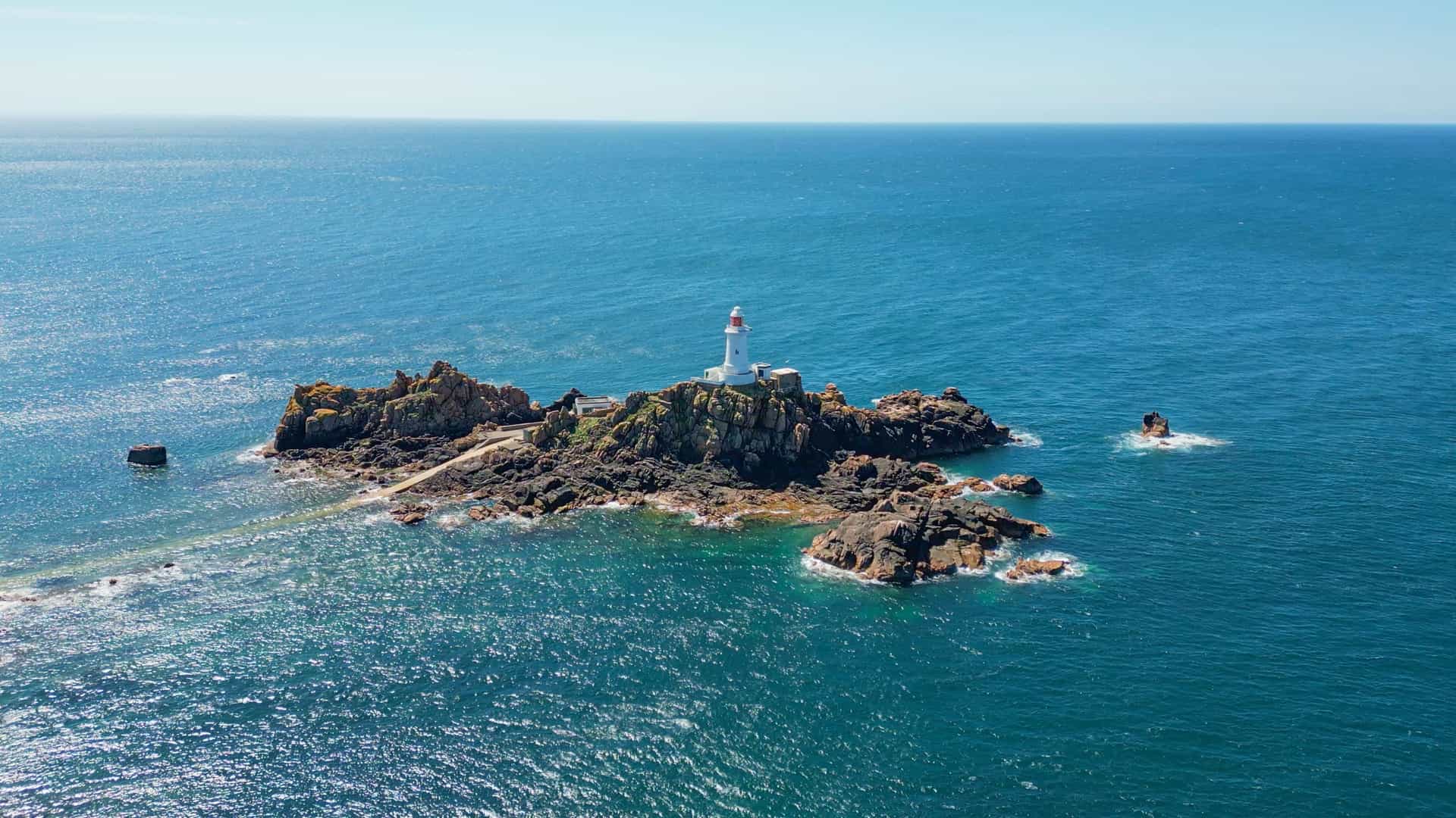 An aerial view of the iconic La Corbière Lighthouse, a popular coastal excursion destination from the St. Helier cruise port in Jersey, Channel Islands. The white lighthouse sits atop a rugged rock formation surrounded by shimmering turquoise waters under a clear, bright blue sky.