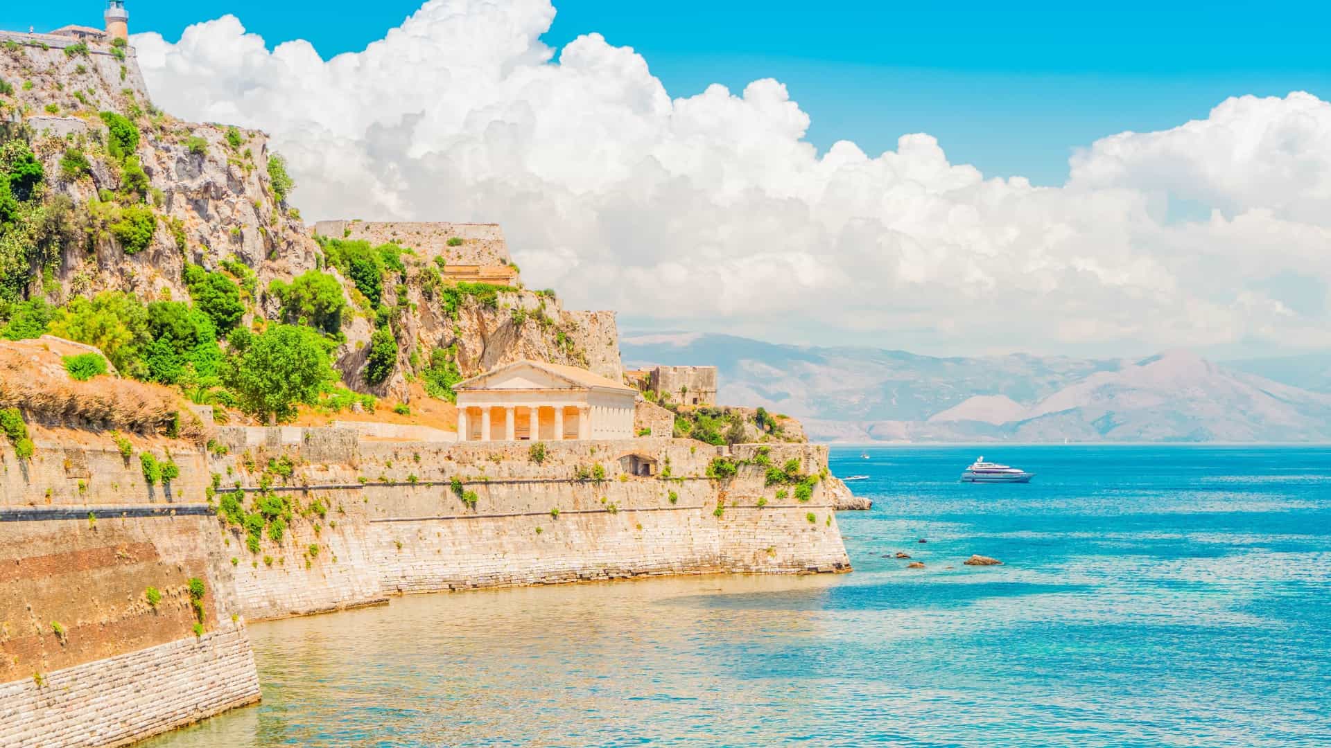 A beautiful coastal view of the Old Fortress of Corfu in Kerkyra, Greece—a prominent shore excursion landmark near the Corfu cruise port. The image highlights the historic stone stone walls and the Doric-style Church of St. George nestled against lush cliffs, looking out over clear blue Ionian waters where a boat cruises in the distance under a partly cloudy sky.