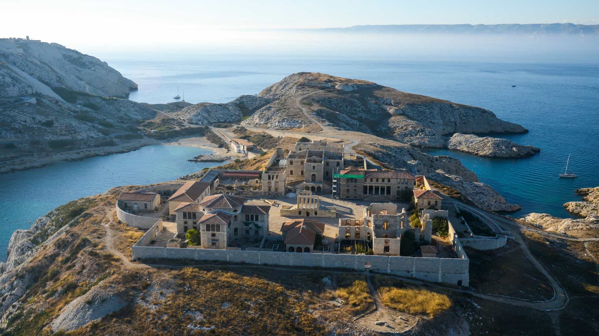 An aerial view of the historic Hôpital Caroline on Ratonneau Island, part of the Frioul archipelago—a popular island excursion destination near the La Joliette cruise port in Marseille, France. The image shows the stone ruins of the former quarantine hospital nestled on a rugged, arid island surrounded by the blue Mediterranean Sea.
