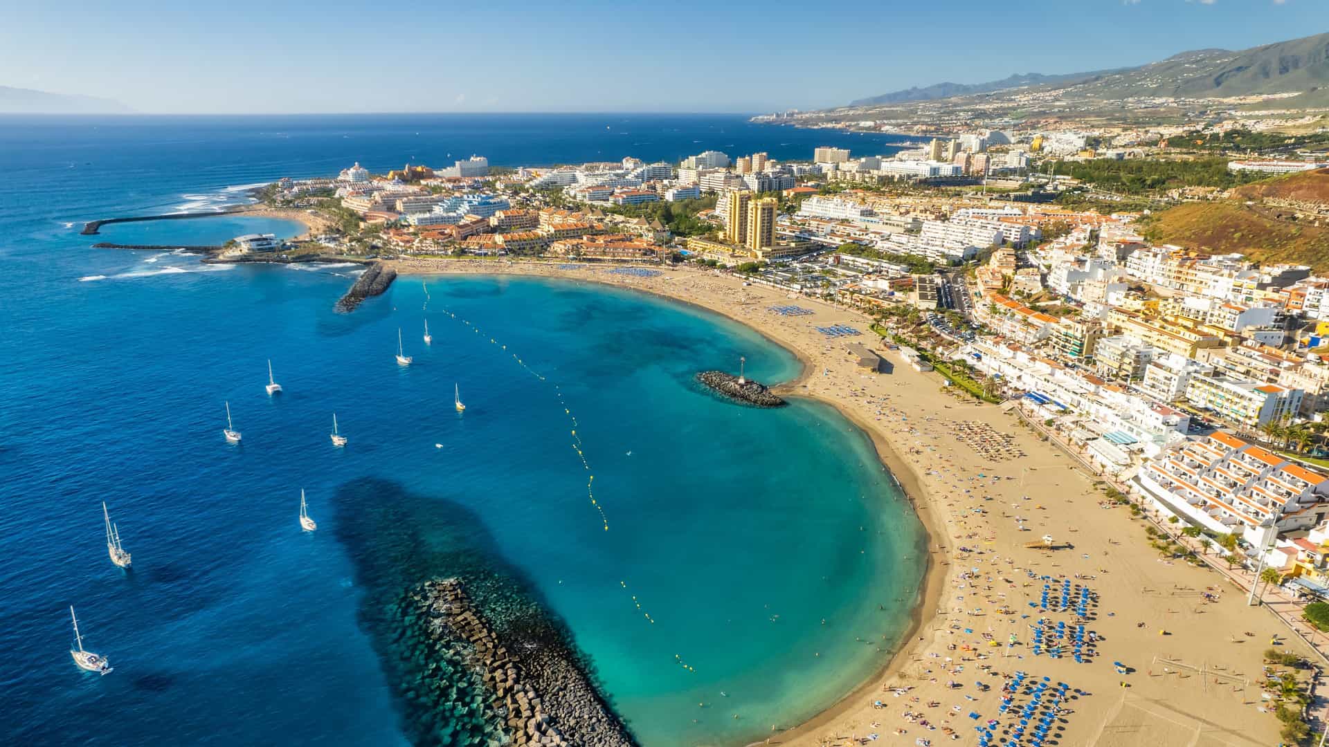 An aerial view of Los Cristianos showing a curved golden‑sand beach, turquoise bay with anchored sailboats, rock breakwaters, and a dense strip of hotels and resorts backed by mountains along the Tenerife coastline.