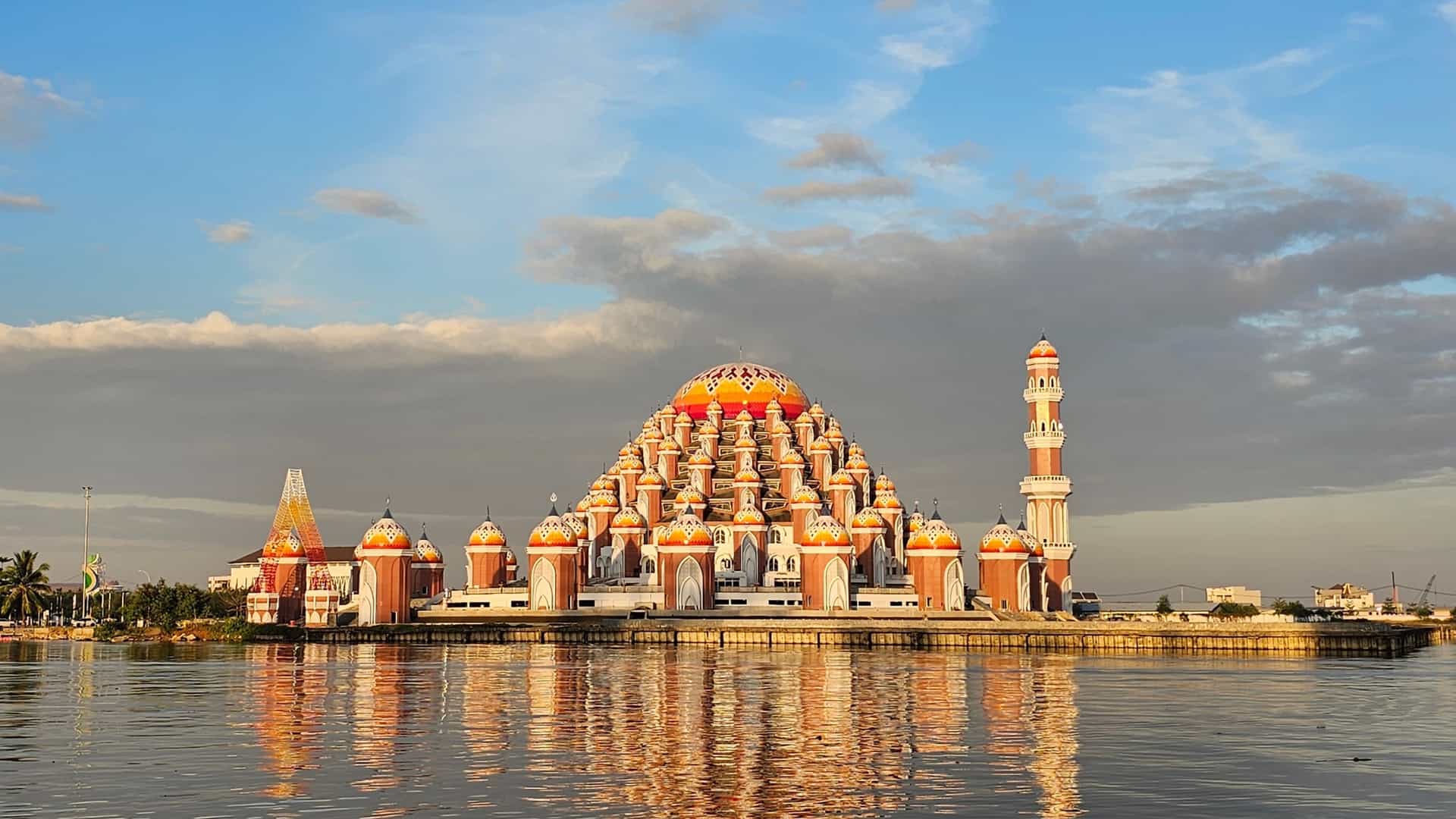 A striking view of the 99 Domes Mosque (Masjid 99 Kubah) on the waterfront of Makassar, South Sulawesi, Indonesia—a landmark cultural destination near the Makassar cruise port. The image showcases the architectural marvel with its uniquely layered, brightly colored orange and red domes reflecting onto the calm water under a partly cloudy sunset sky.