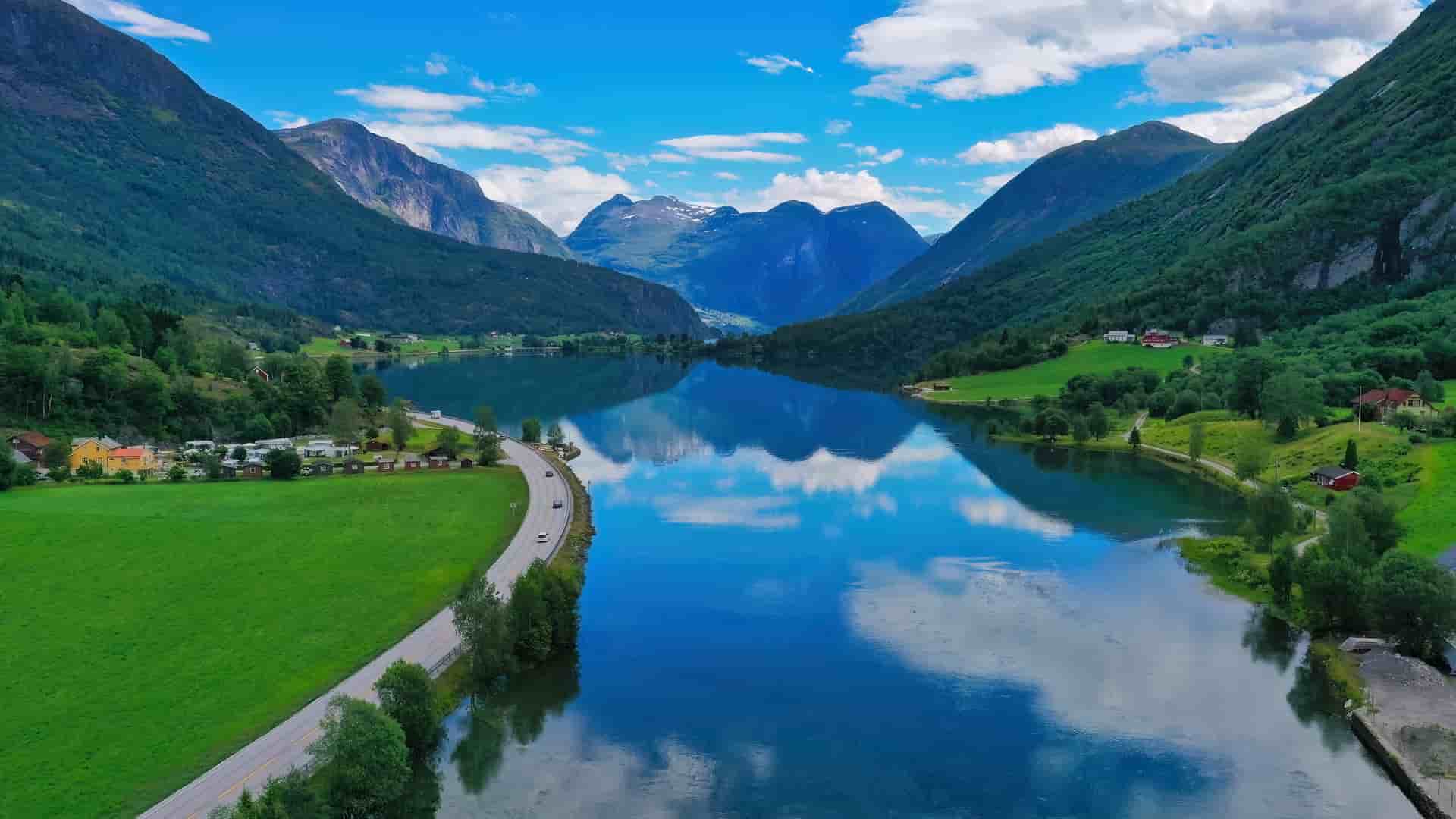 A breathtaking aerial view of a glassy, deep blue fjord or lake in the Nordfjord region (Nordfjordland) of Norway, a world-renowned cruise destination. The image showcases steep, lush green mountains perfectly mirrored on the water's surface, flanked by a scenic lakeside road, scattered rural houses, and bright green fields under a vibrant blue sky with fluffy white clouds.