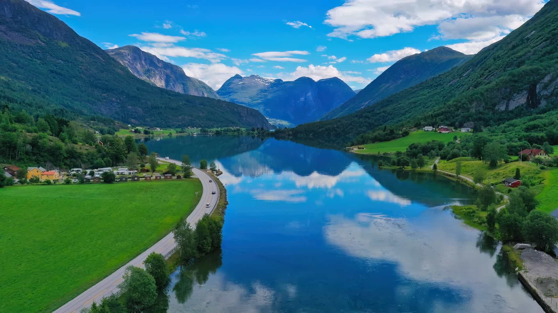 A breathtaking aerial view of a glassy, deep blue fjord or lake in the Nordfjord region (Nordfjordland) of Norway, a world-renowned cruise destination. The image showcases steep, lush green mountains perfectly mirrored on the water's surface, flanked by a scenic lakeside road, scattered rural houses, and bright green fields under a vibrant blue sky with fluffy white clouds.