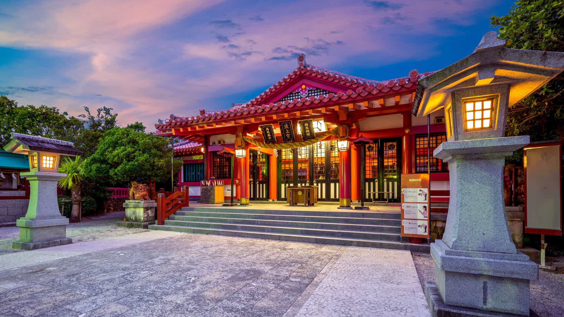 A serene twilight view of the historic Naminoue Shrine in Okinawa, Japan—a popular cultural shore excursion near the Motobu and Naha cruise ports. The image highlights the traditional Okinawan architecture of the shrine's main pavilion, illuminated by glowing stone lanterns on a stone-paved courtyard under a vibrant sunset sky with hues of purple, orange, and blue.