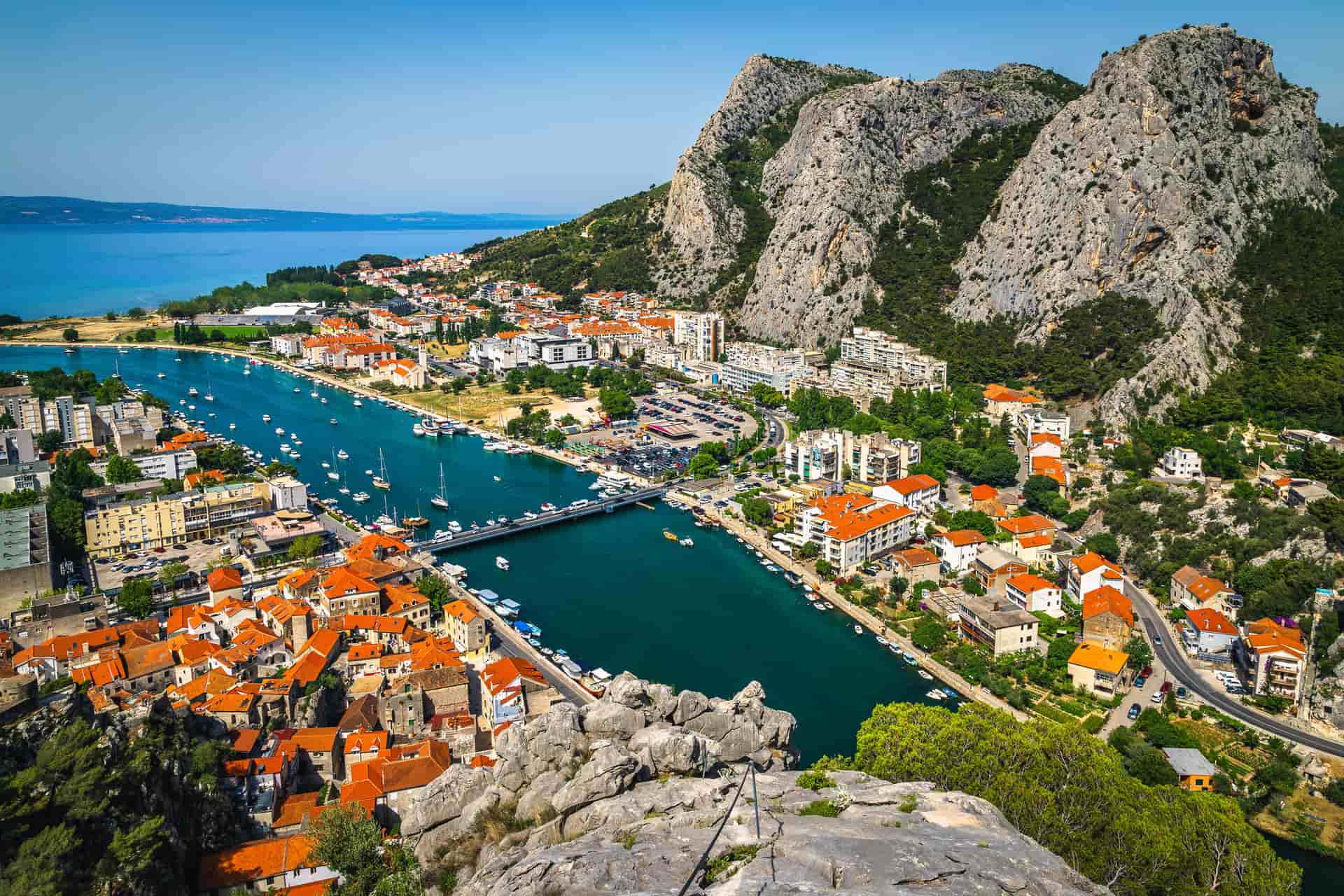An aerial panorama of the coastal town of Omiš, Croatia—a breathtaking destination located southeast of the Split cruise port. The image showcases the Cetina River cutting through a dramatic canyon of massive, rugged limestone cliffs before meeting the turquoise Adriatic Sea, with the red-tiled roofs of the historic town, docked boats, and a local bridge visible below.