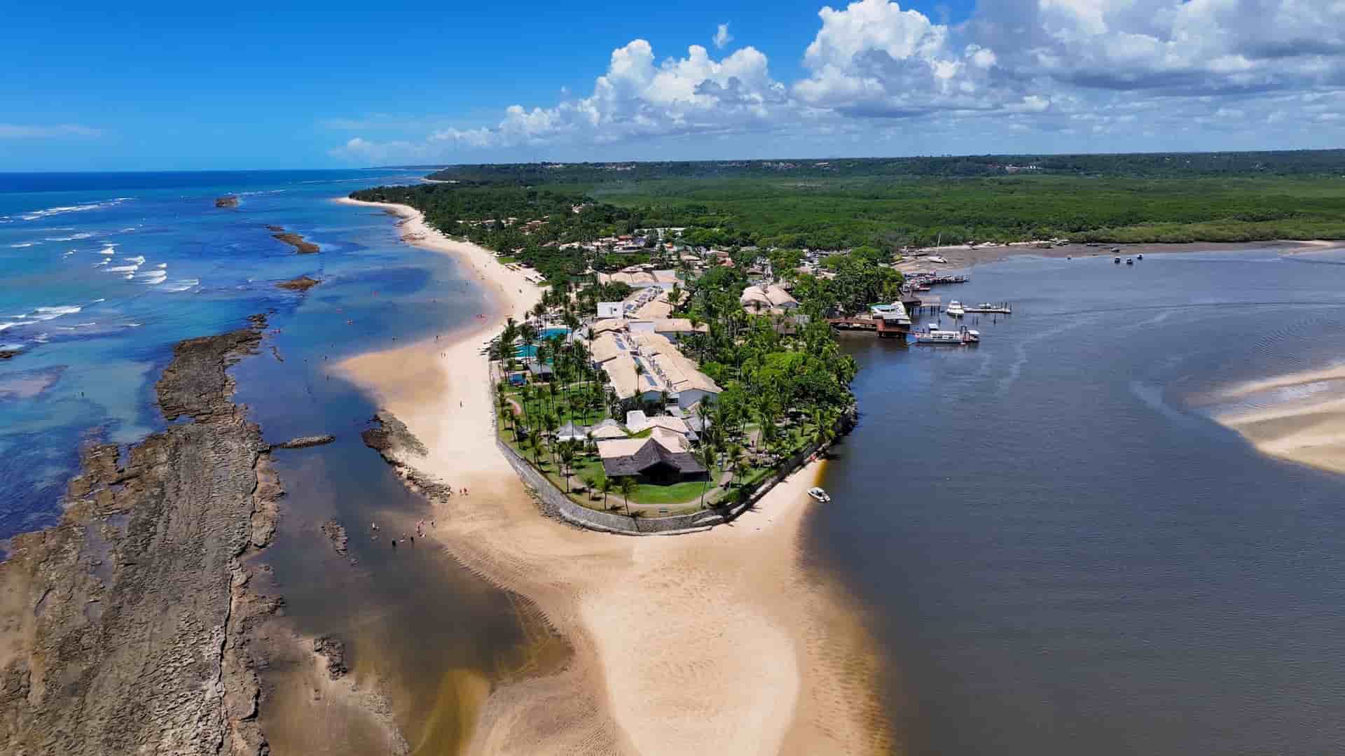 An aerial view of the tropical coastline at Porto Seguro in Bahia, Brazil—a historic South American cruise destination. The image showcases a long, sandy peninsula lined with palm trees and beachside resorts, bordered on one side by a shallow reef in the blue Atlantic Ocean and on the other by a calm river estuary and dense green mangroves under a bright, scattered cloud sky.